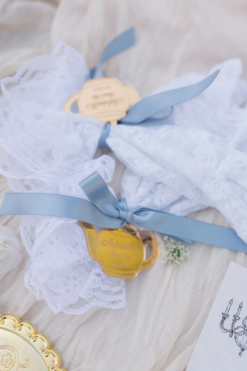 Close-up of a lace wedding throw with blue ribbon, a gold keychain with the text "Antoinette's Bridal Tea, Est. 2015," and a small sprig of white flowers, all on a white fabric surface.