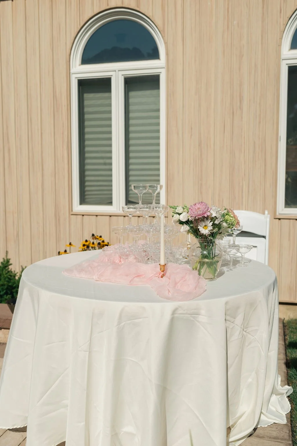 Decorative table set with champagne glasses, a pink flower arrangement, and a white candle, outdoors against a wooden building with arched windows.