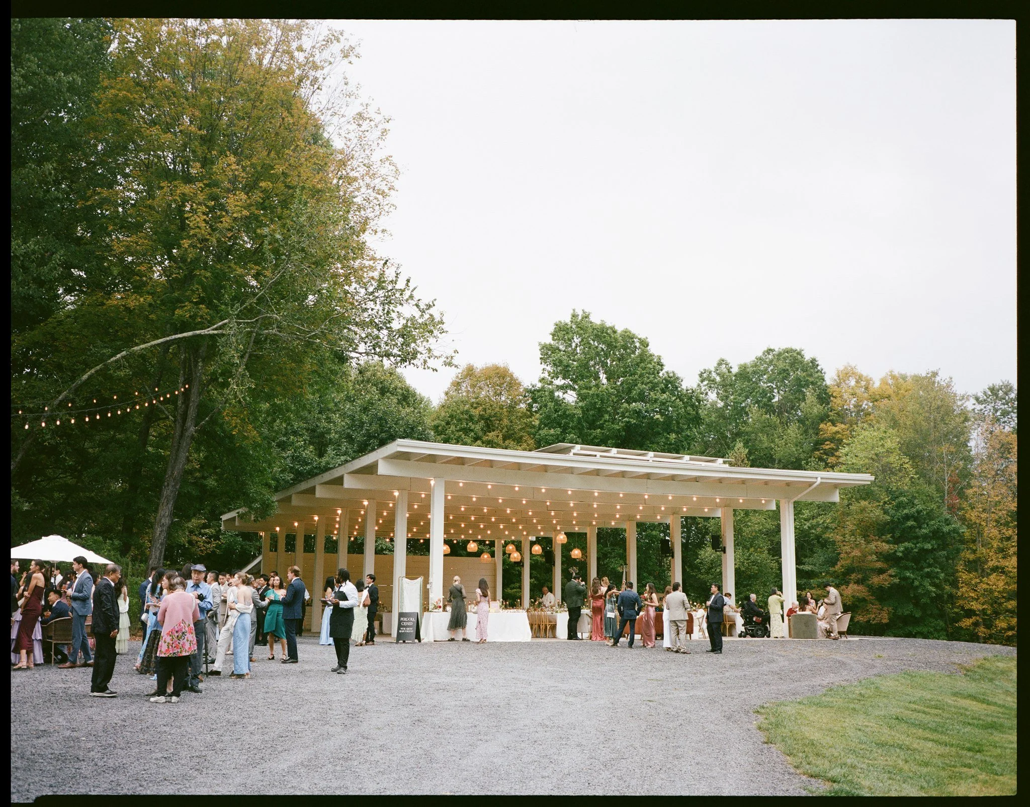 Outdoor wedding reception with guests gathered around a decorated pavilion and long buffet table, surrounded by trees with fall foliage.