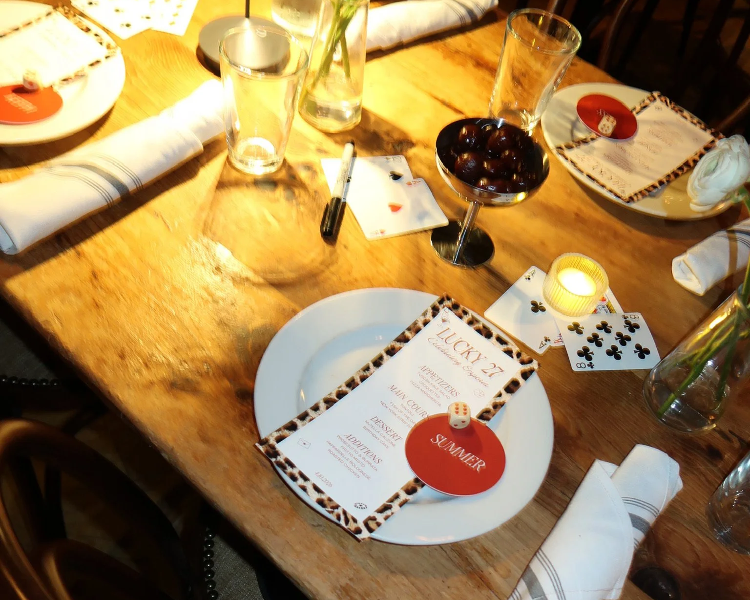 A wooden dining table set for a celebration with menus, playing cards, a small lit candle, glasses, a bowl of cherries, and decorative items.