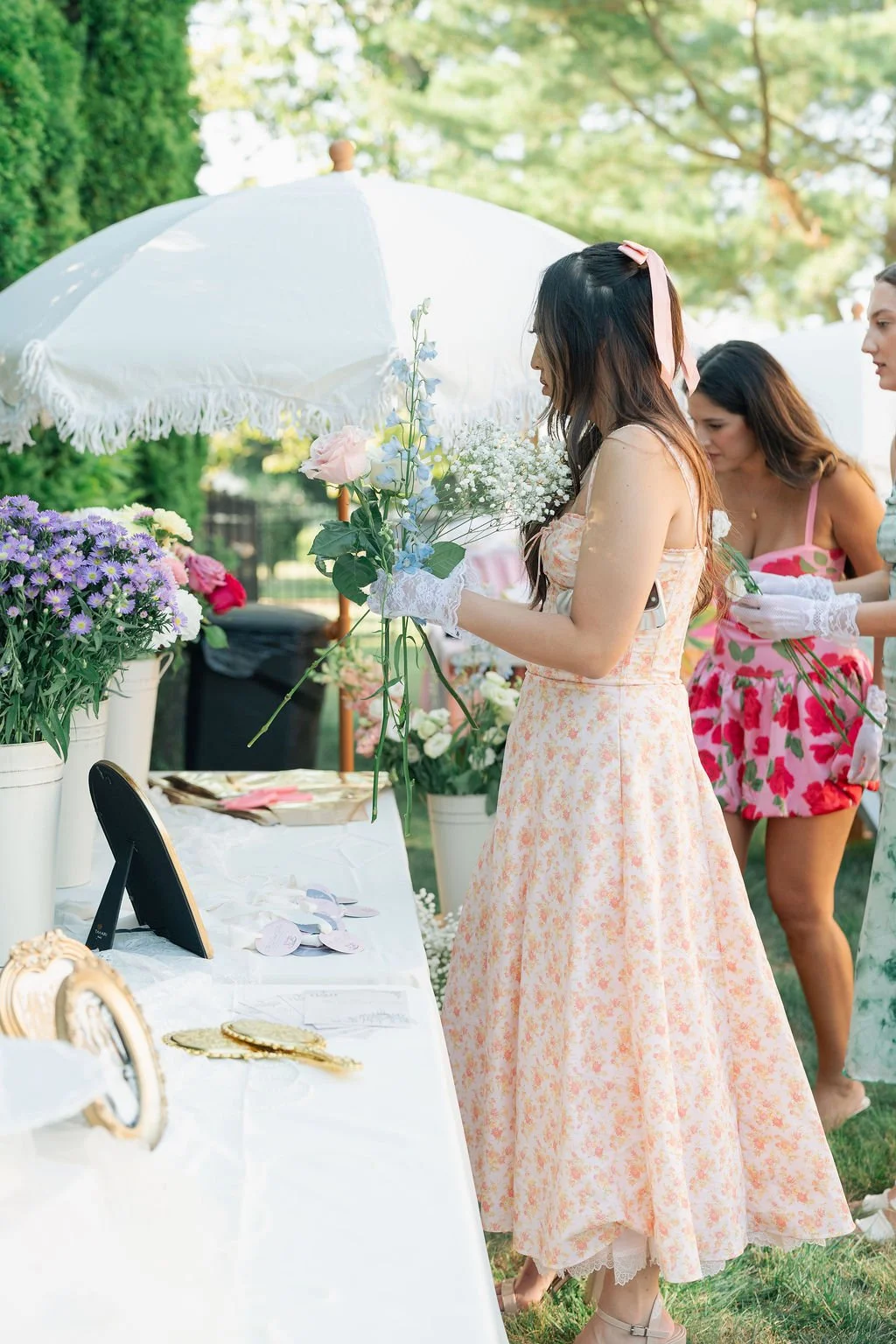 Women preparing floral arrangements at an outdoor garden party, with flowers, a white parasol, and decorative items on a table.