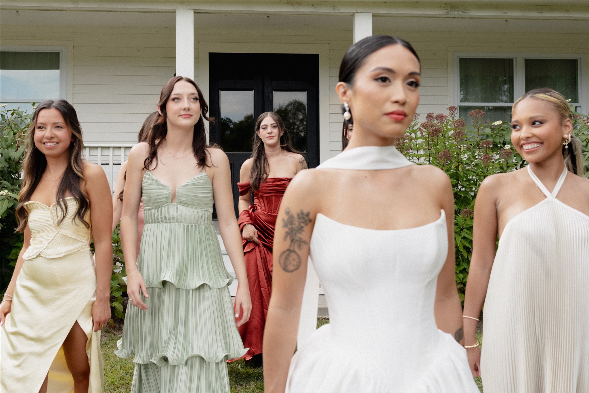 A group of women standing outdoors in front of a house, dressed in formal dresses, with some smiling and looking in different directions.