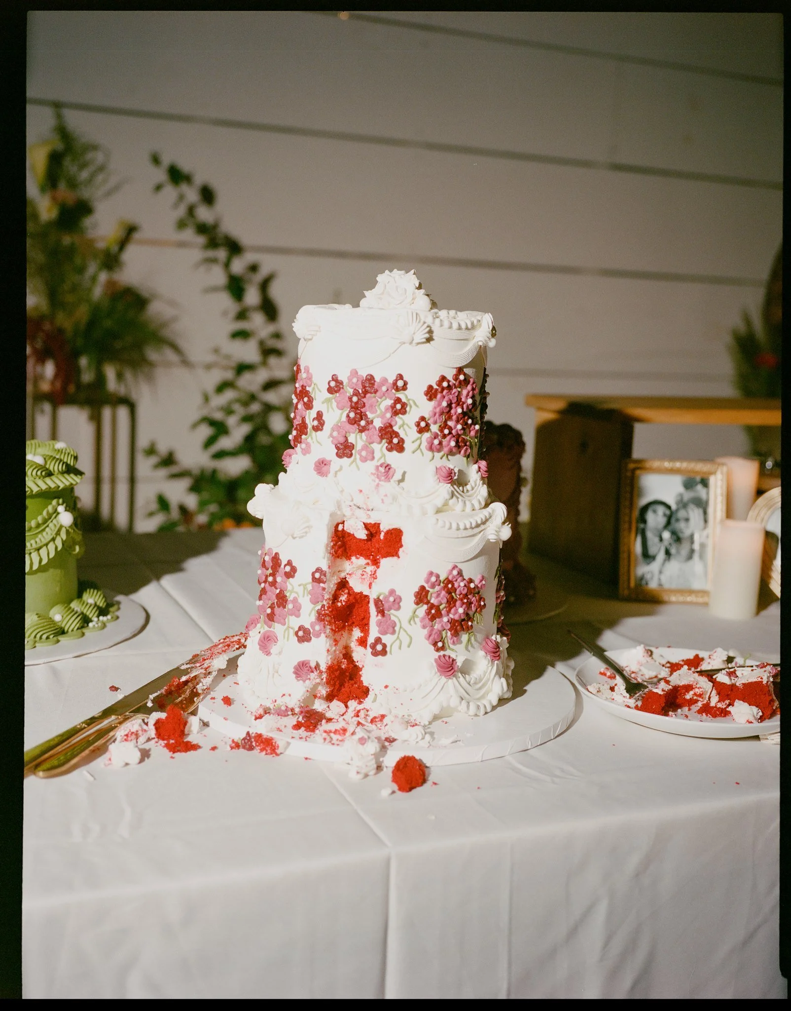 A multi-tiered white wedding cake decorated with red and pink flowers and green leaves, placed on a white tablecloth, with red crumbles and frosting scattered around and a cake knife and spatula nearby.