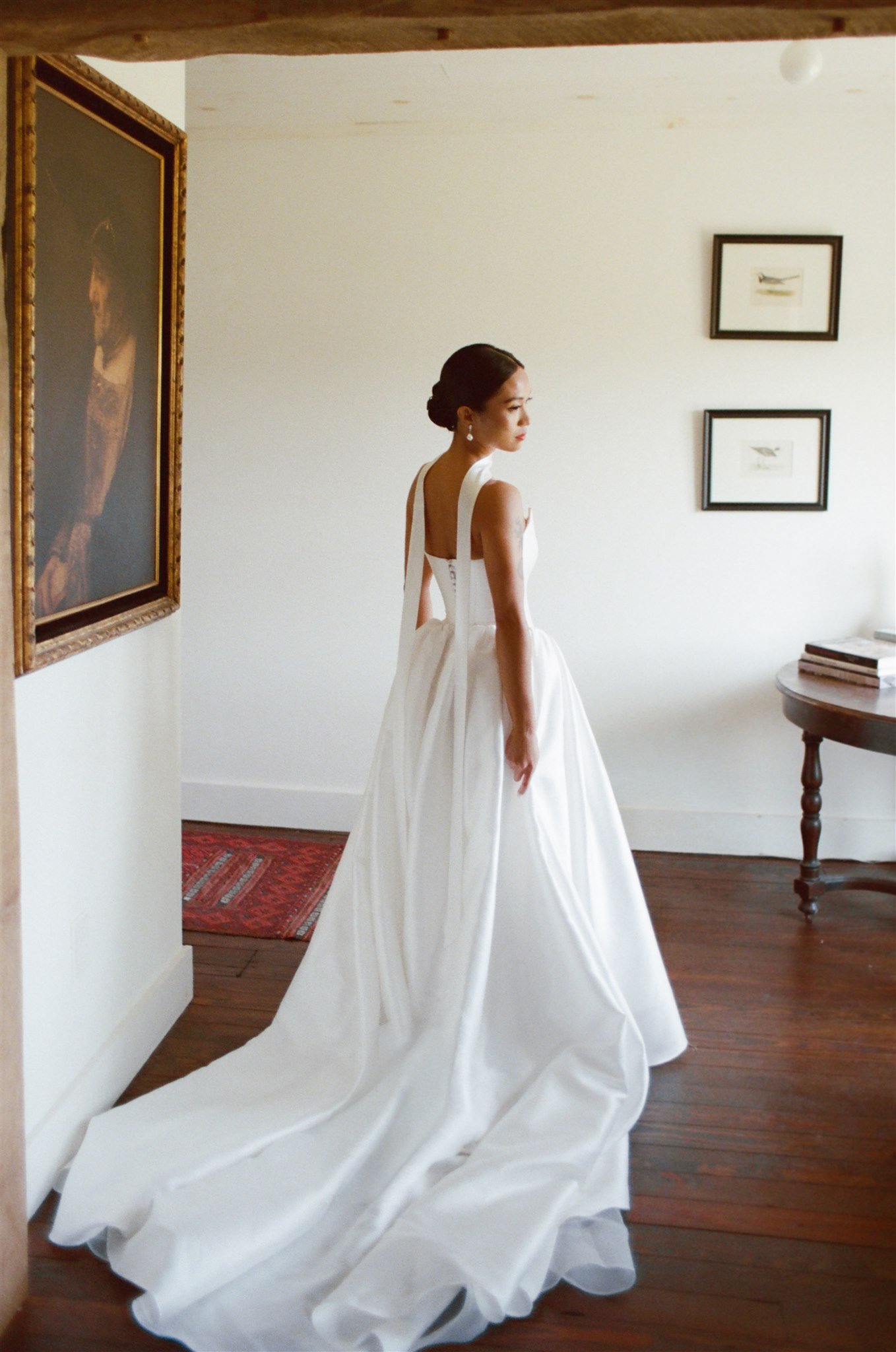 A woman in a white wedding gown standing in a room with wooden floor, framed pictures on the wall, and a small table with books.
