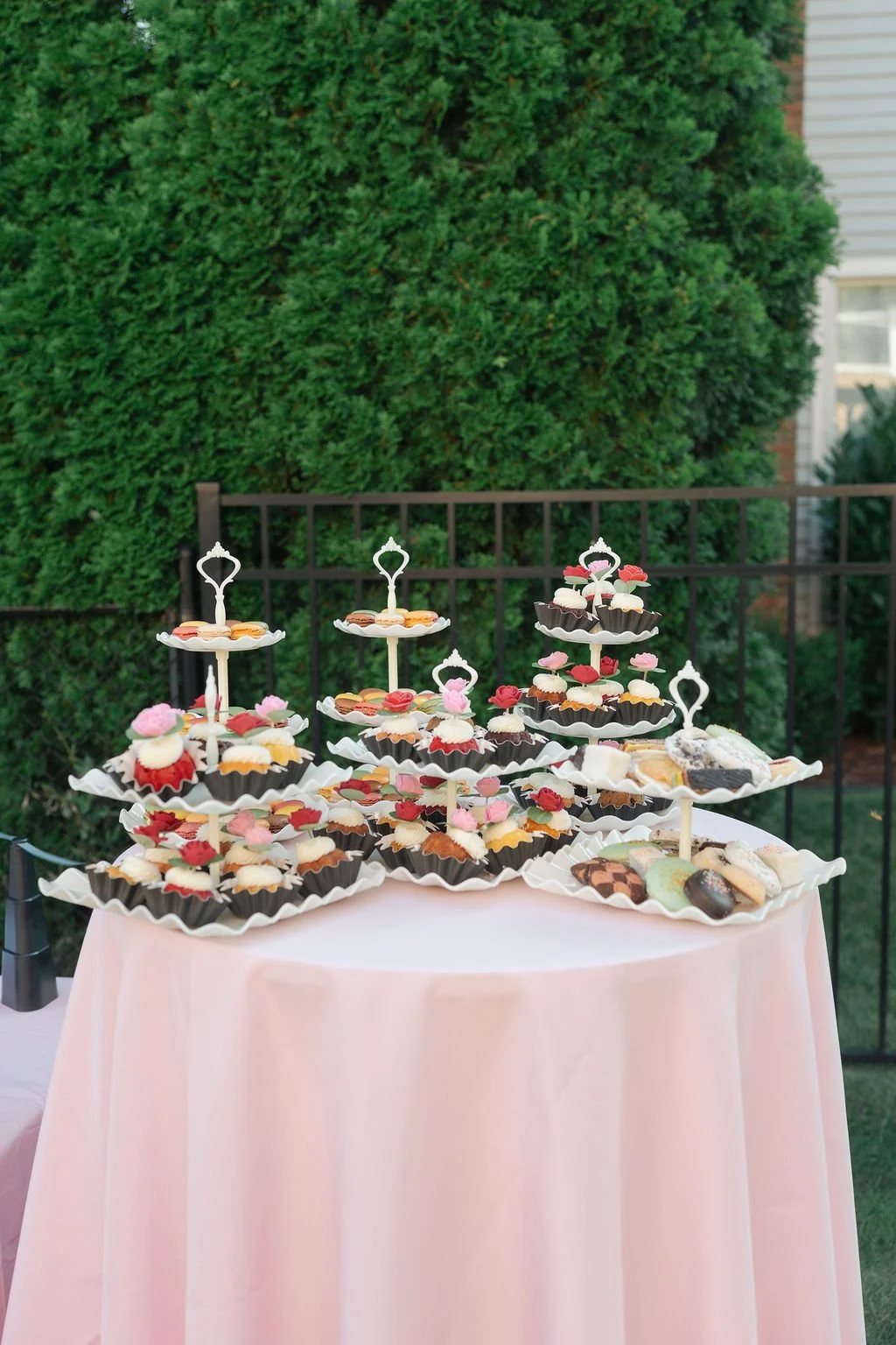 Tiered dessert stand with assorted cupcakes and cookies on a pink tablecloth outdoors, with green shrubbery and a fence in the background.