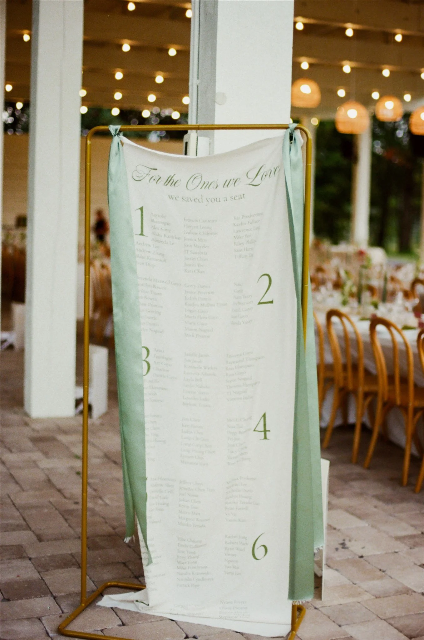 A moth-colored fabric wedding seating chart with numbered sections and guest names hanging from a gold metal frame at an outdoor event, with chairs arranged in the background and string lights overhead.