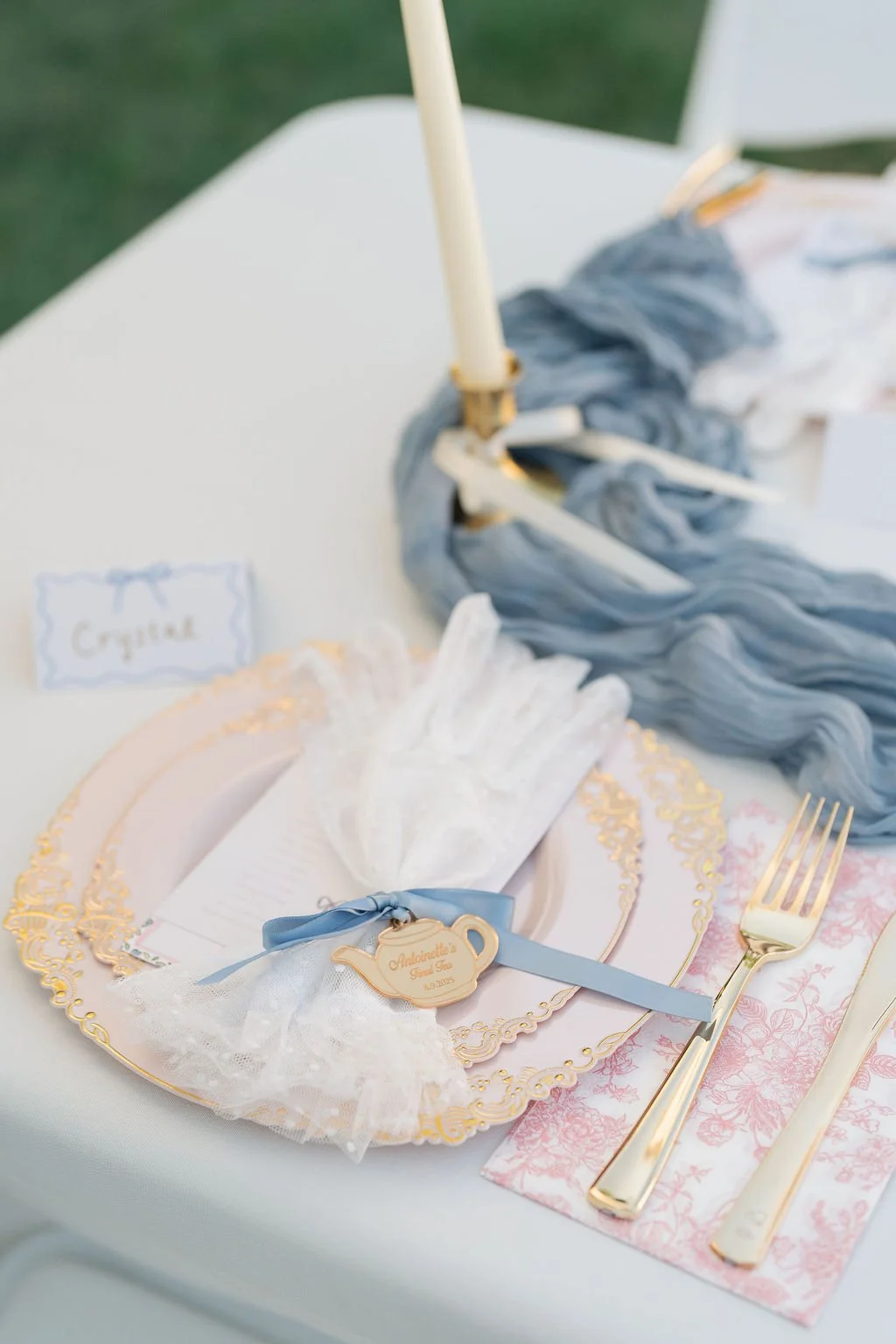 Elegant table setting with gold utensils, a lace napkin, and a teapot-shaped tag on a blue ribbon, part of a themed decor with a large gray fabric ruffle and a white taper candle in the background.