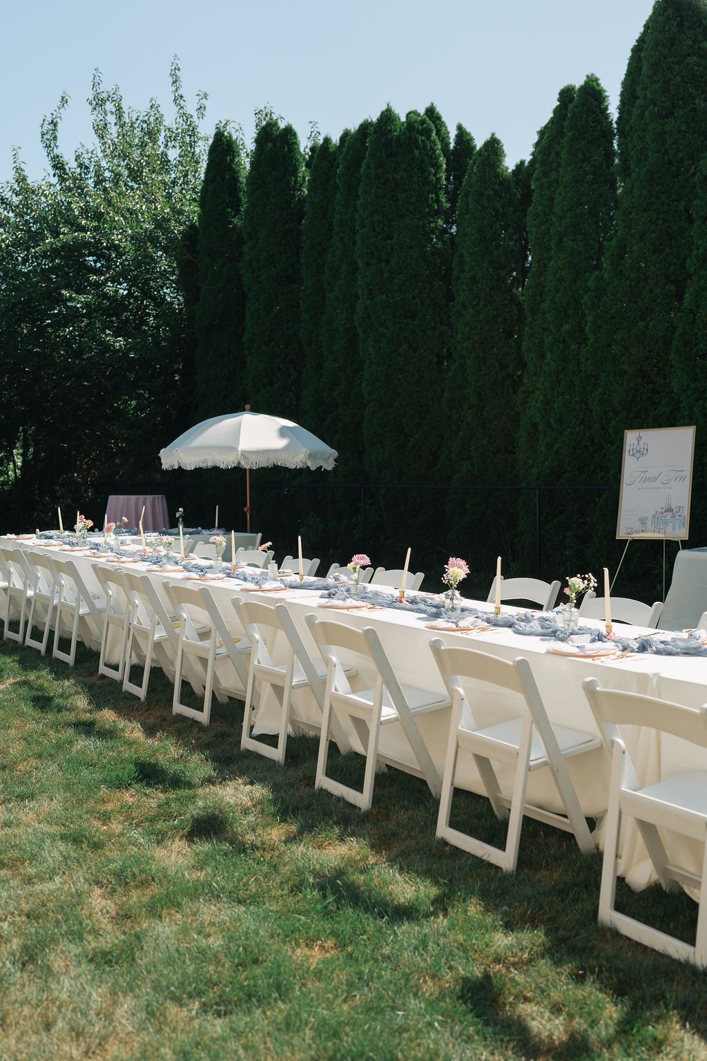 Long outdoor table set for a celebration with white chairs, floral centerpieces, and candles, under a large white umbrella, on a grassy lawn with tall green trees in the background.