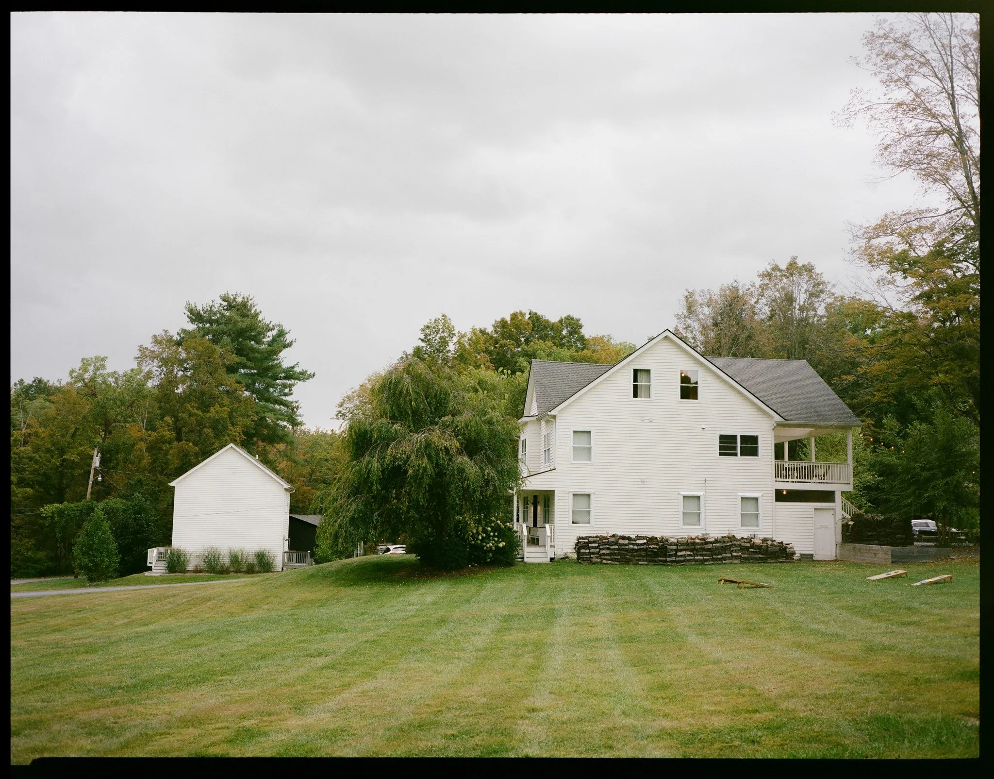 A white house with gray roof and multiple windows, surrounded by green trees, and a well-maintained lawn with a pile of firewood and some small objects in the yard, under a cloudy sky.