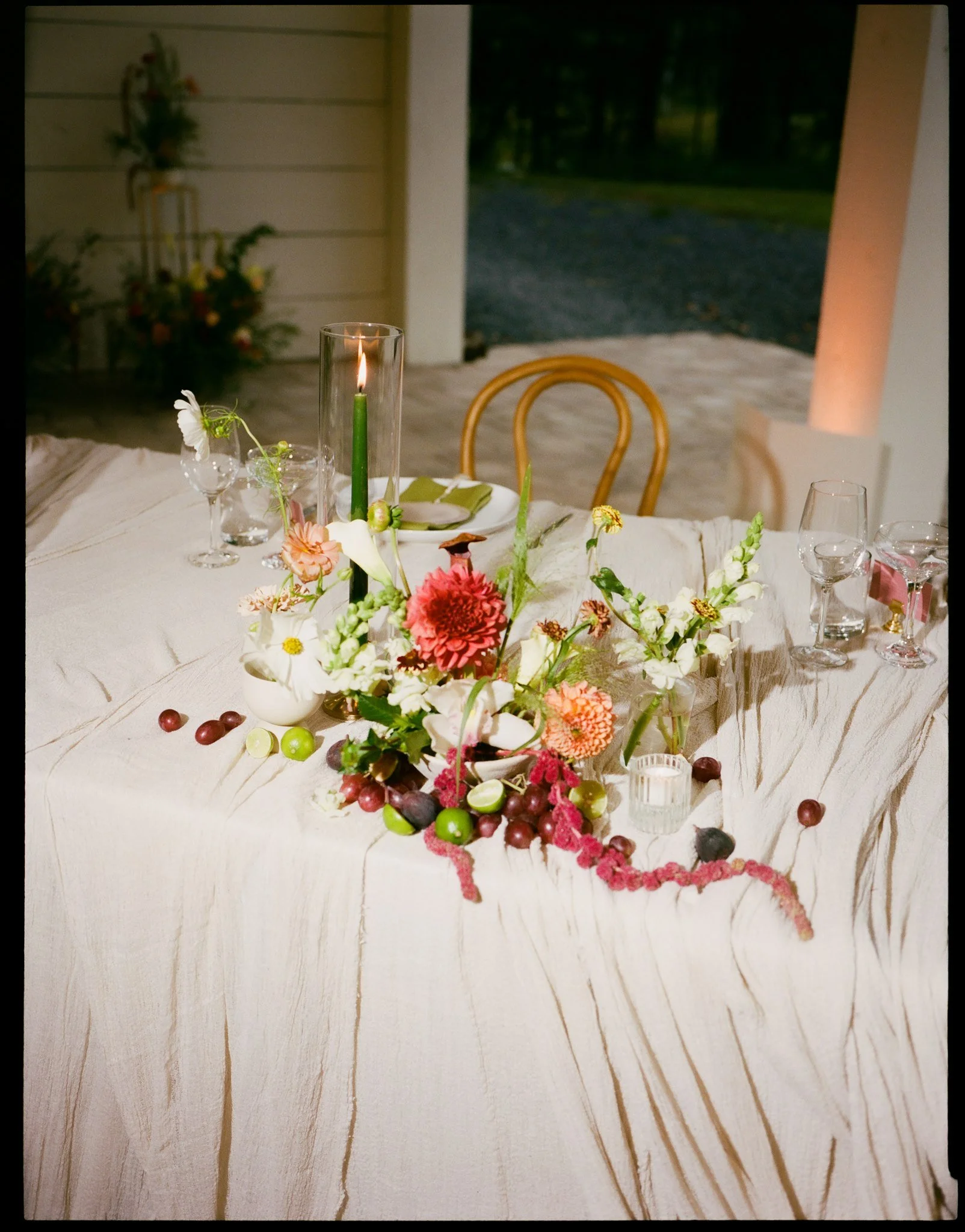 A decorated table with a floral centerpiece, a tall candle, and scattered berries. Outside, a porch and trees are visible.