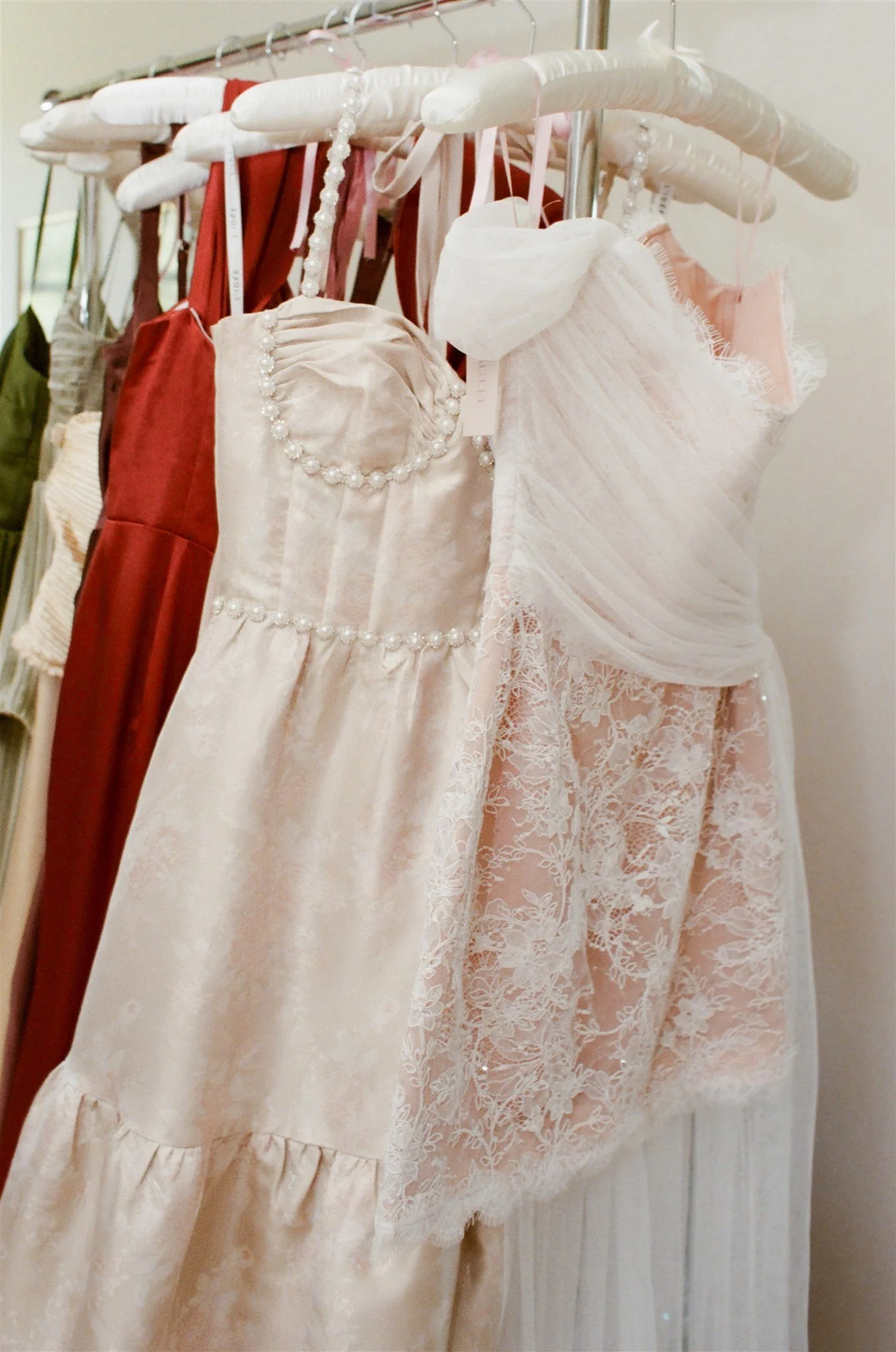A row of dresses hanging on padded hangers, featuring various shades of pink, cream, and red, with lace, ruffles, and pearl embellishments.