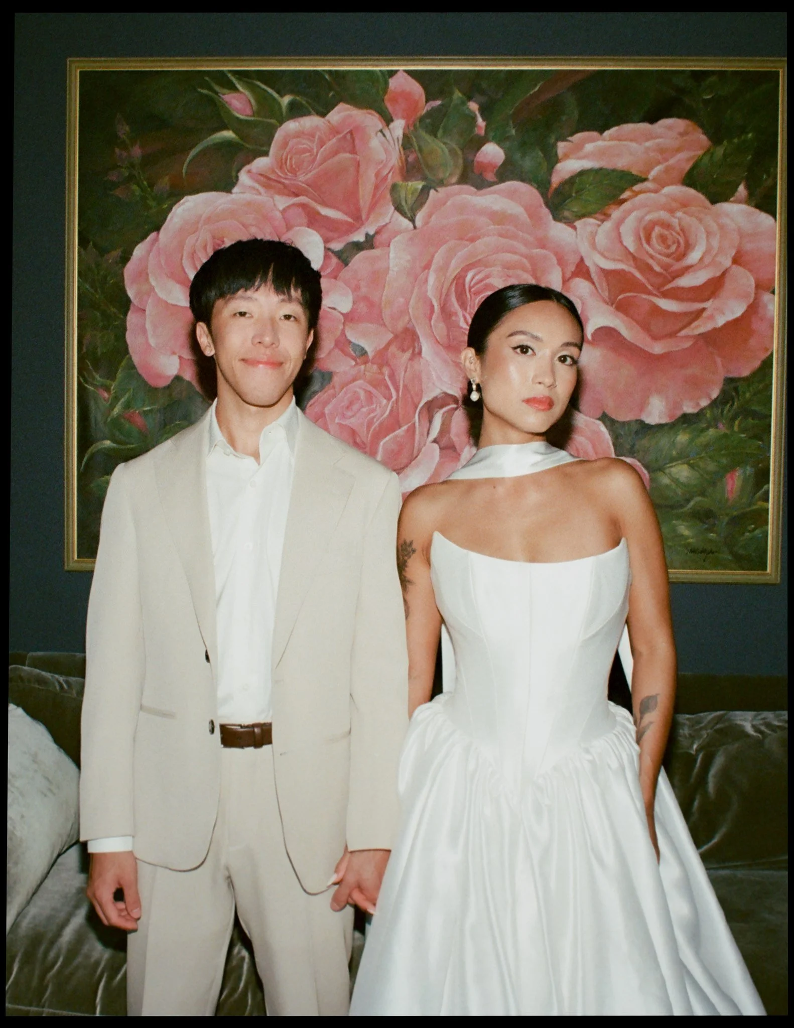 A man and a woman dressed in white standing in front of a large painting of pink roses. The man is smiling with his eyes closed, wearing a white suit and shirt. The woman has a serious expression, with dark hair pulled back, wearing a white strapless