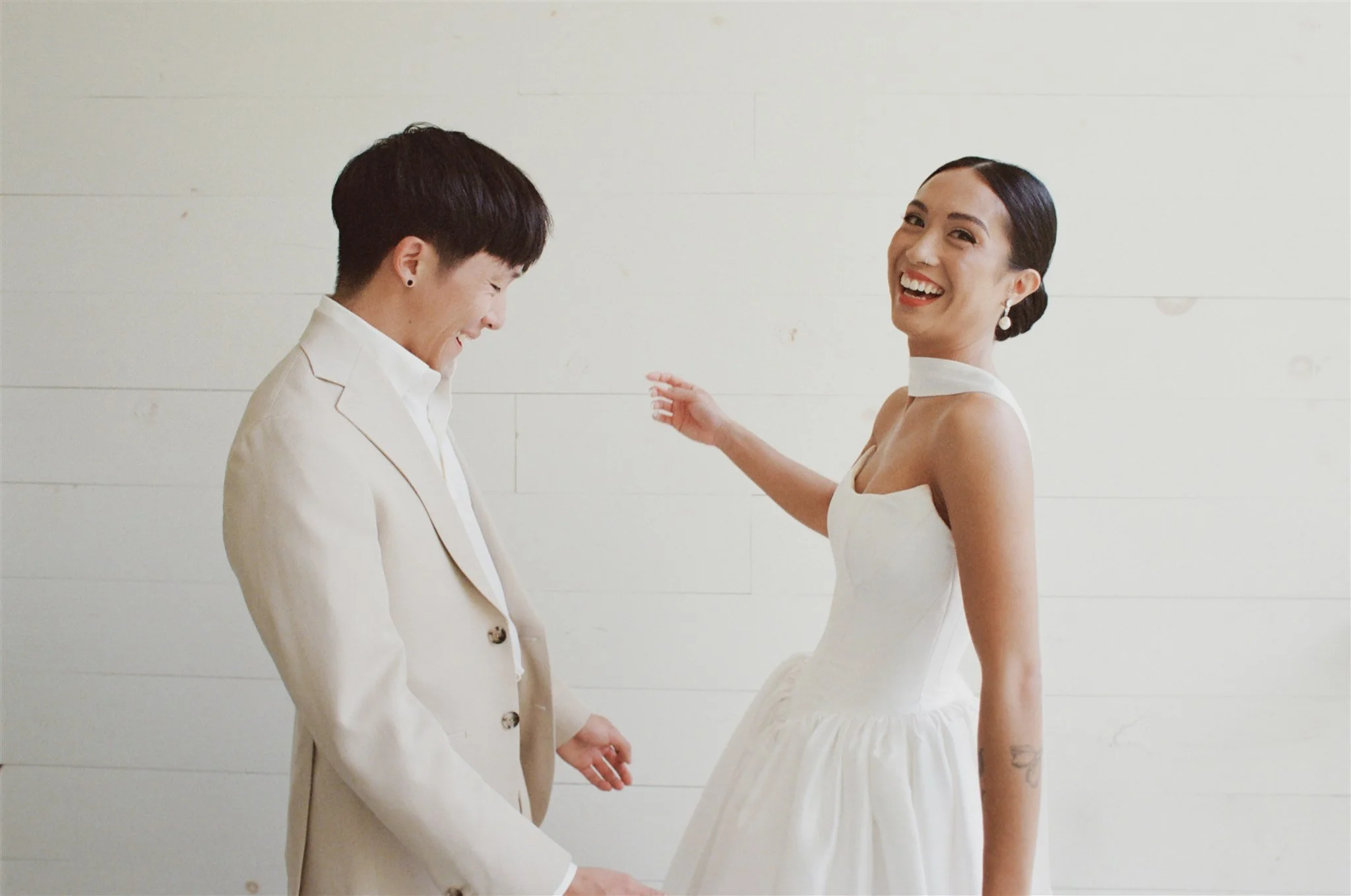 A man and woman in formal attire smiling and holding hands against a white wall background.