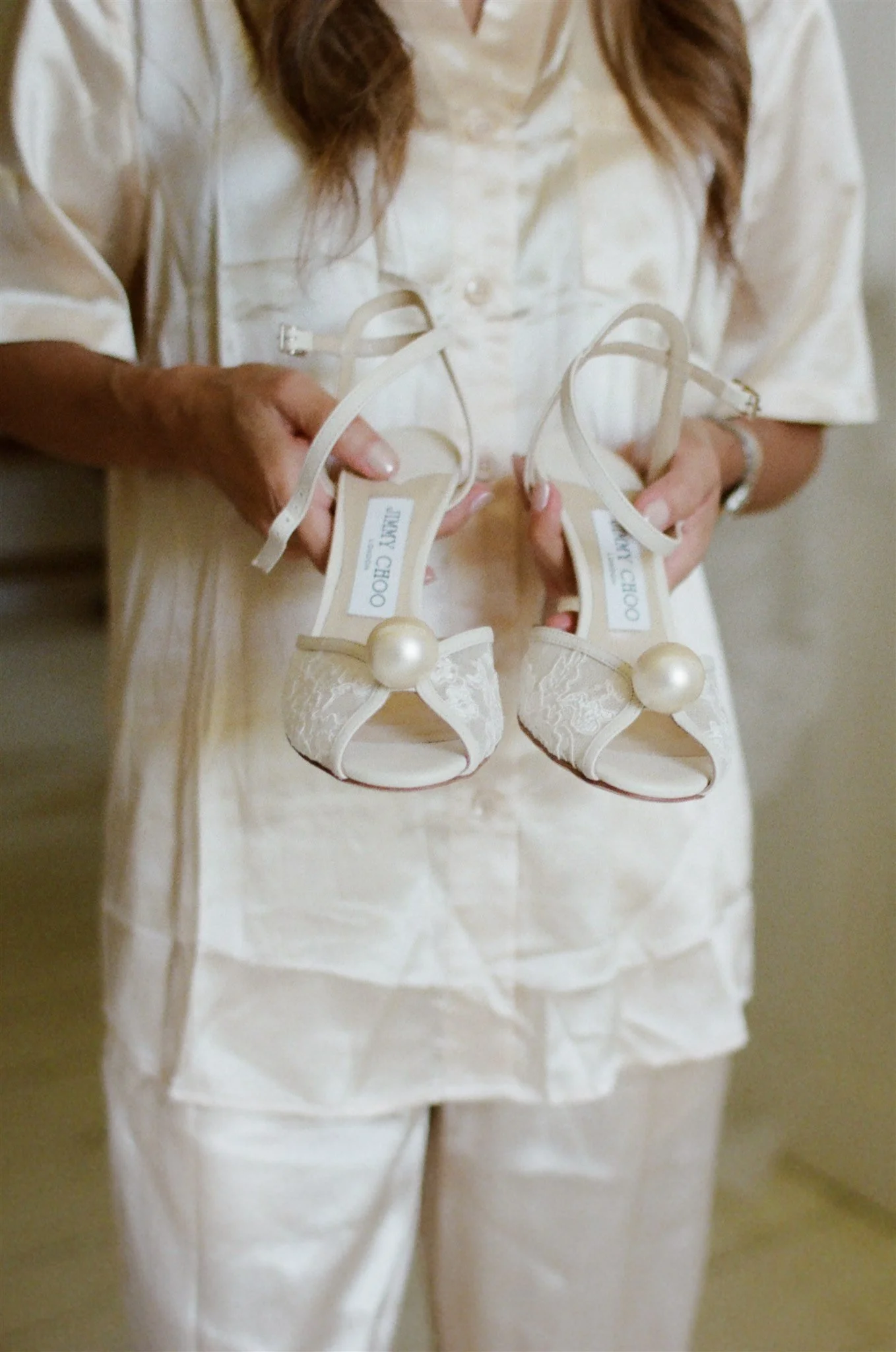 A person holding a pair of white high-heeled shoes with pearl accents.