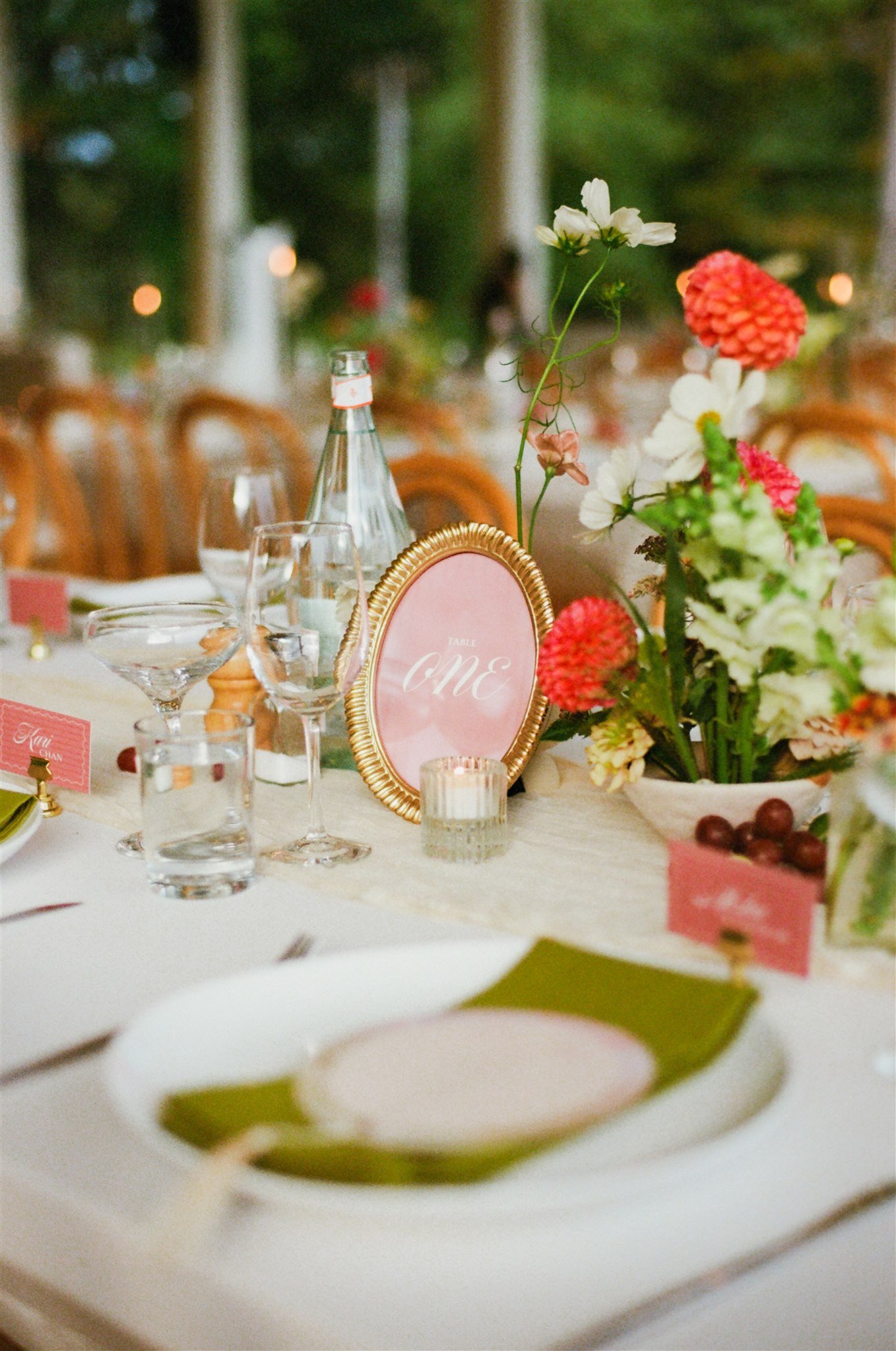 A close-up of a decorated table for a celebration or wedding, featuring floral centerpieces with white and red flowers, glassware, a pink frame with the word "ONE," and a candle.