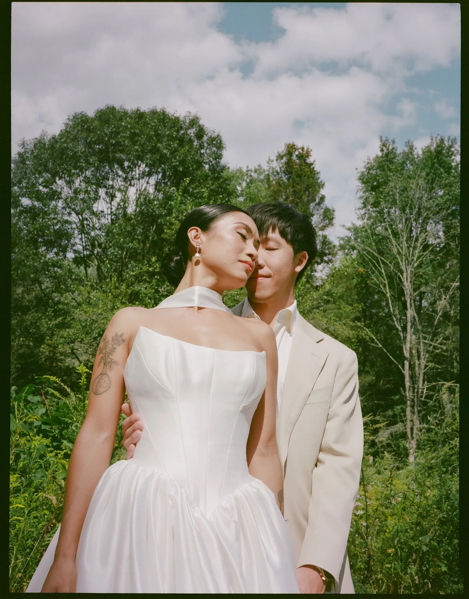 A couple is embracing outdoors among green trees, with her eyes closed and him smiling gently, both dressed in formal white attire.