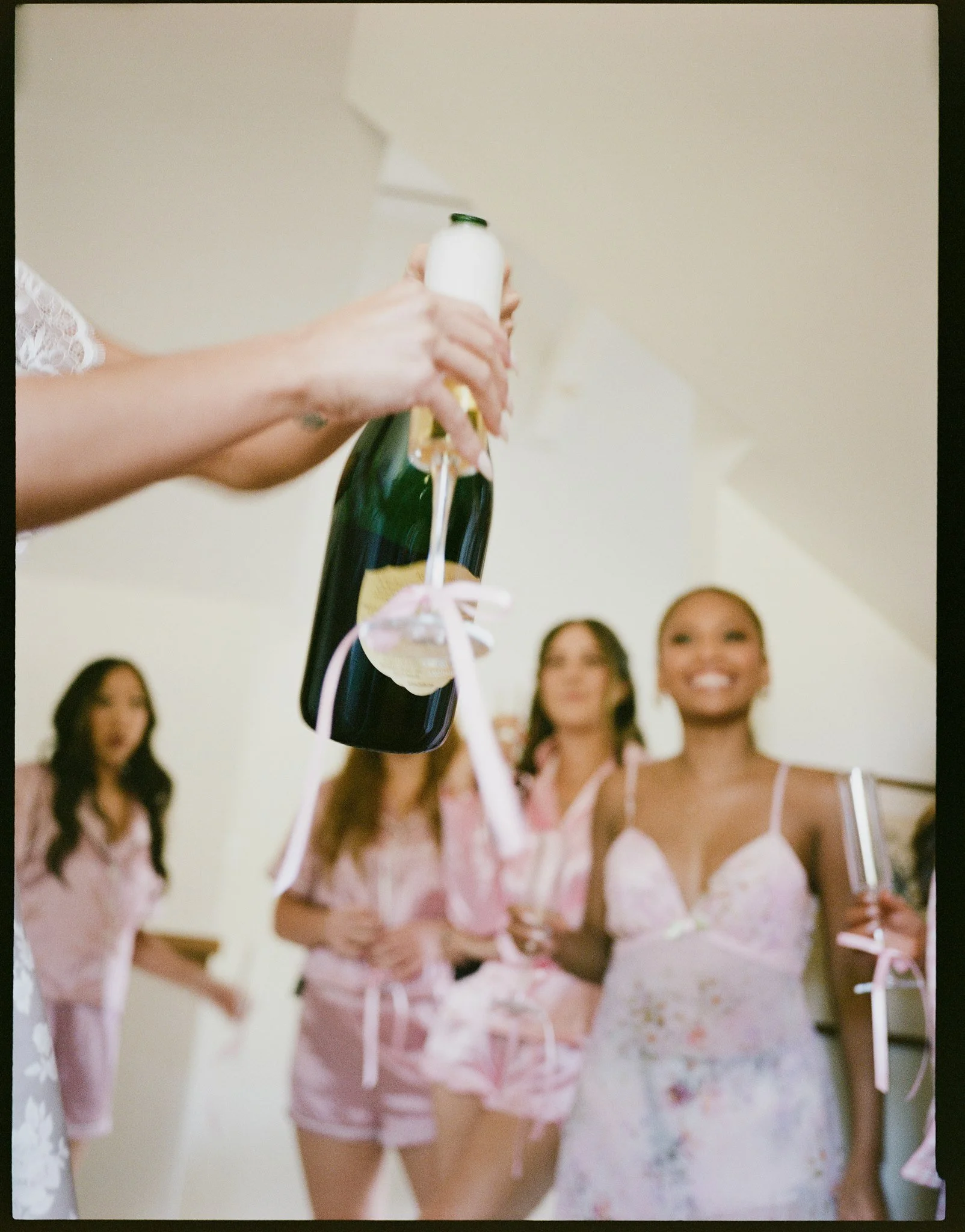 A person holding a champagne bottle with a white ribbon, a label, and foam spilling from the top, while four women in pajamas smile and pose in the background.