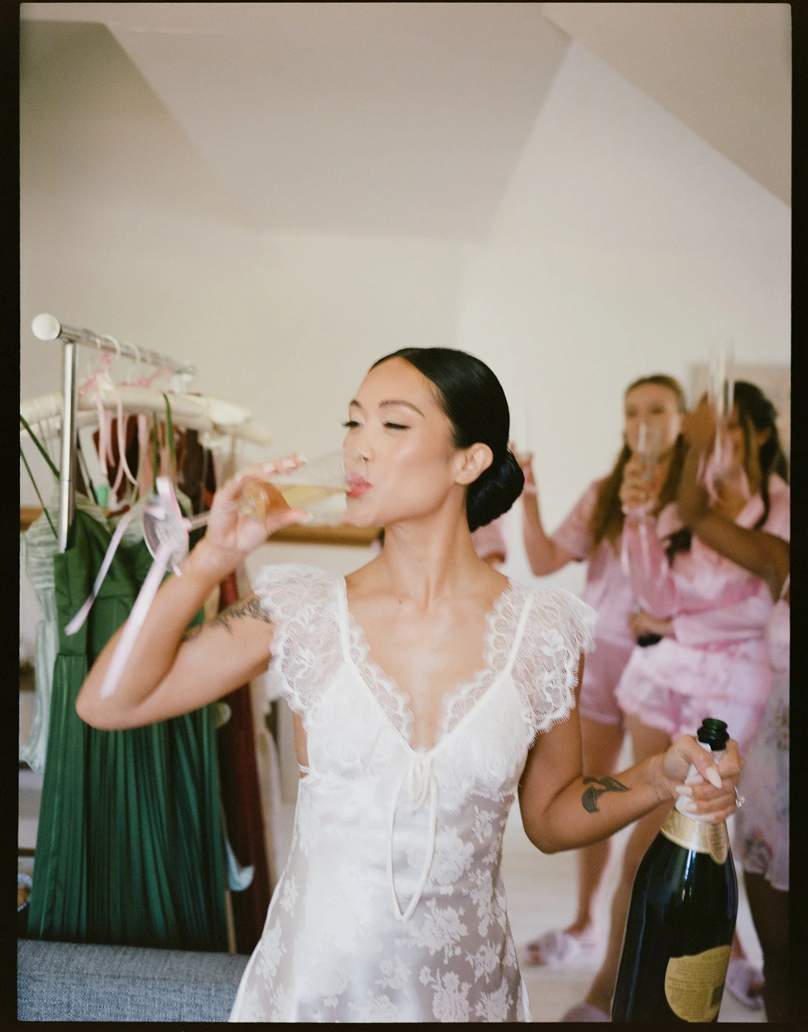 A woman in a white lace dress drinking from a champagne flute at a celebration, with bridesmaids in pink robes in the background.