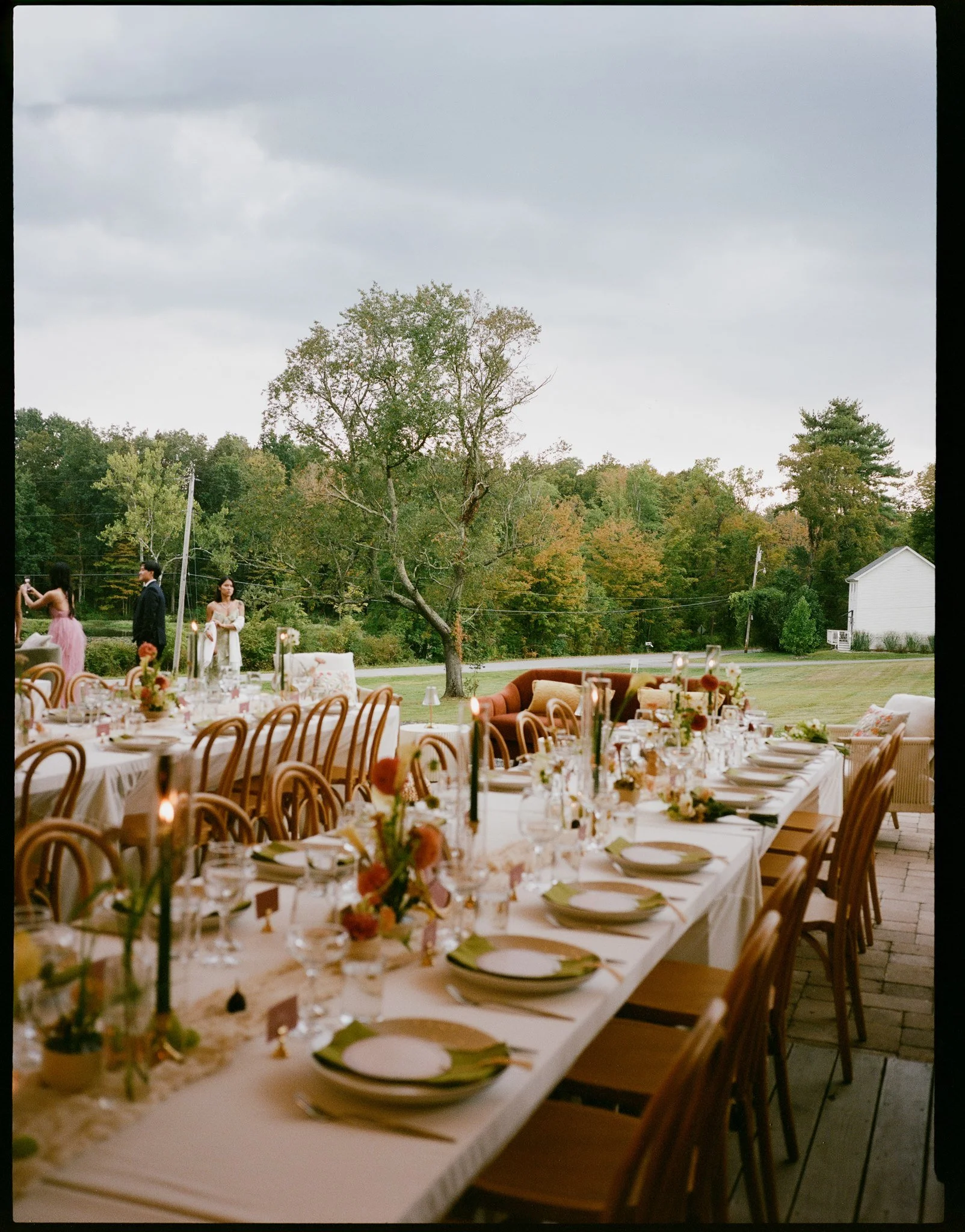 Long dining table set up outdoors with beige chairs, floral centerpieces, candles, and place settings, under a cloudy sky, with trees and a house in the background.