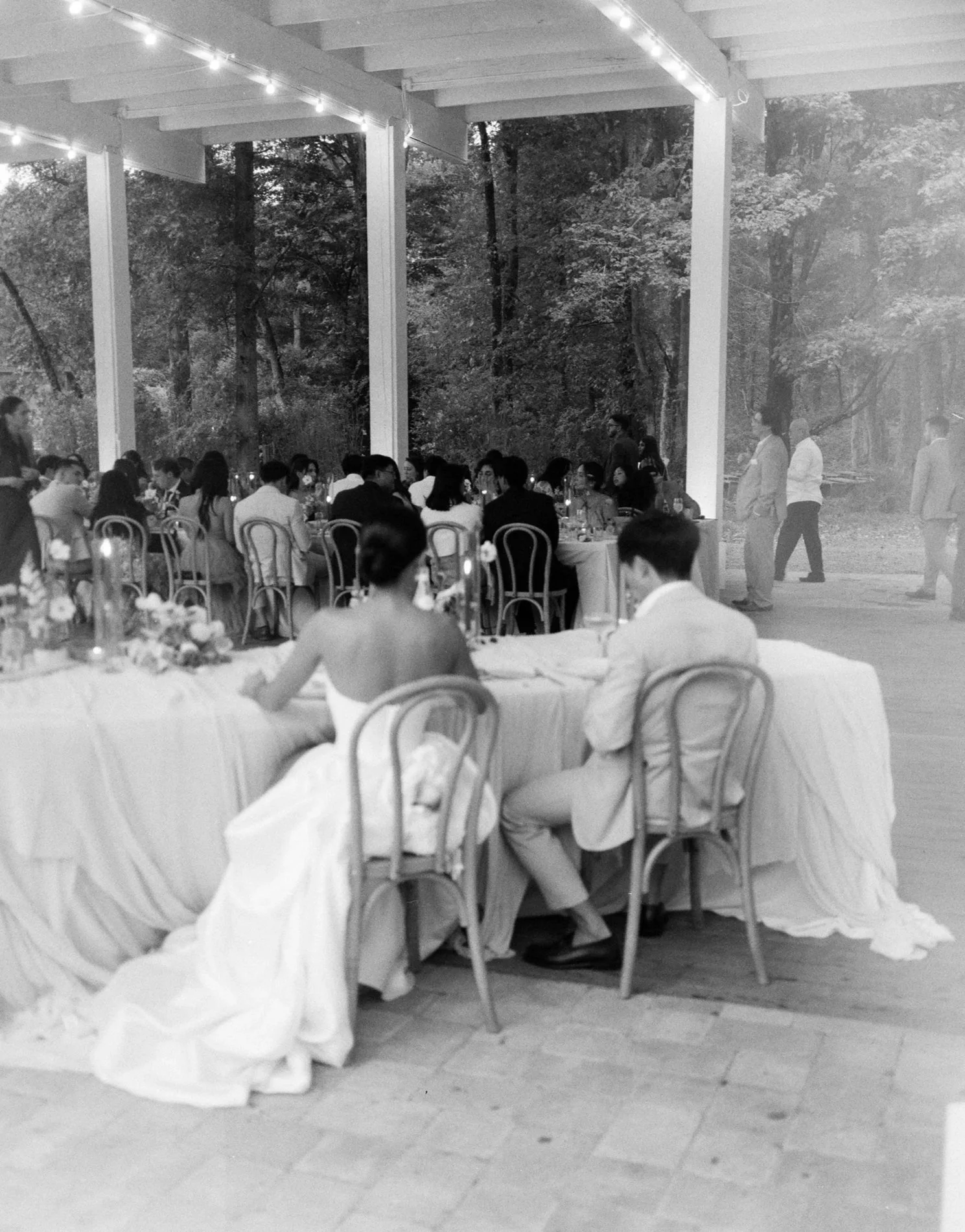 Black and white photo of a wedding reception with a large group of people seated at round tables under a patio with string lights. Two women are in the foreground, one with short hair and a strapless wedding dress, and the other with long hair, both facing away from the camera.