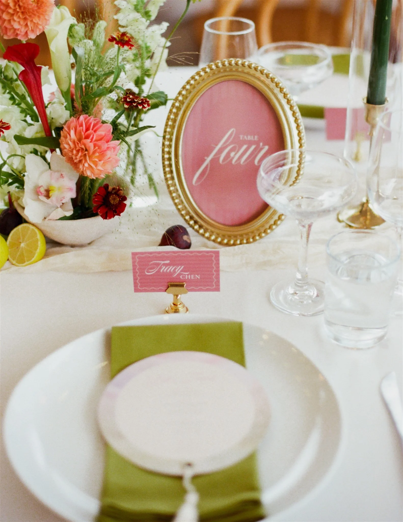 A table setting with a centerpiece of pink and white flowers, a pink sign with gold trim that reads 'Table Four,' and empty white bowls surrounded by clear glassware and green napkins.