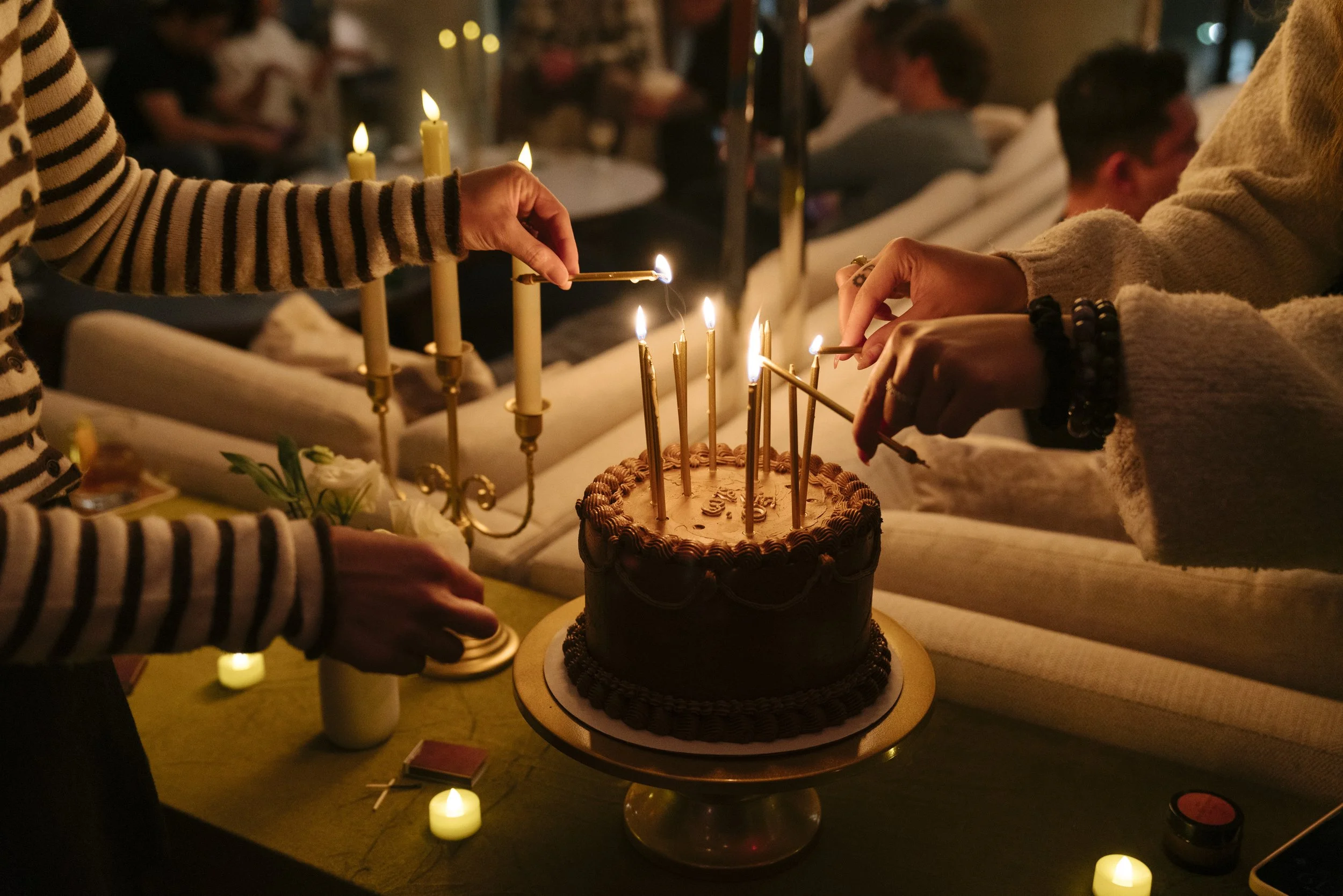 People gather around a chocolate birthday cake with lit candles, as one person lights a candle with a match in a cozy, dimly lit setting.
