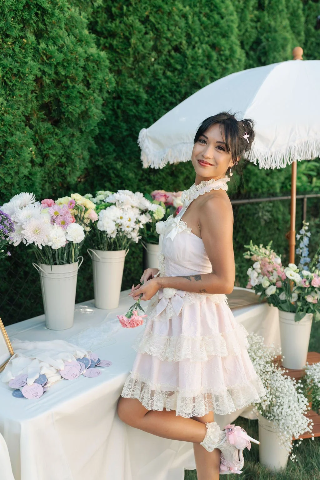 Young woman in a white lace dress with pink accents stands outdoors with various colorful flowers in white vases and a white tablecloth behind her, holding a pink flower, with a white parasol and green hedge in the background.