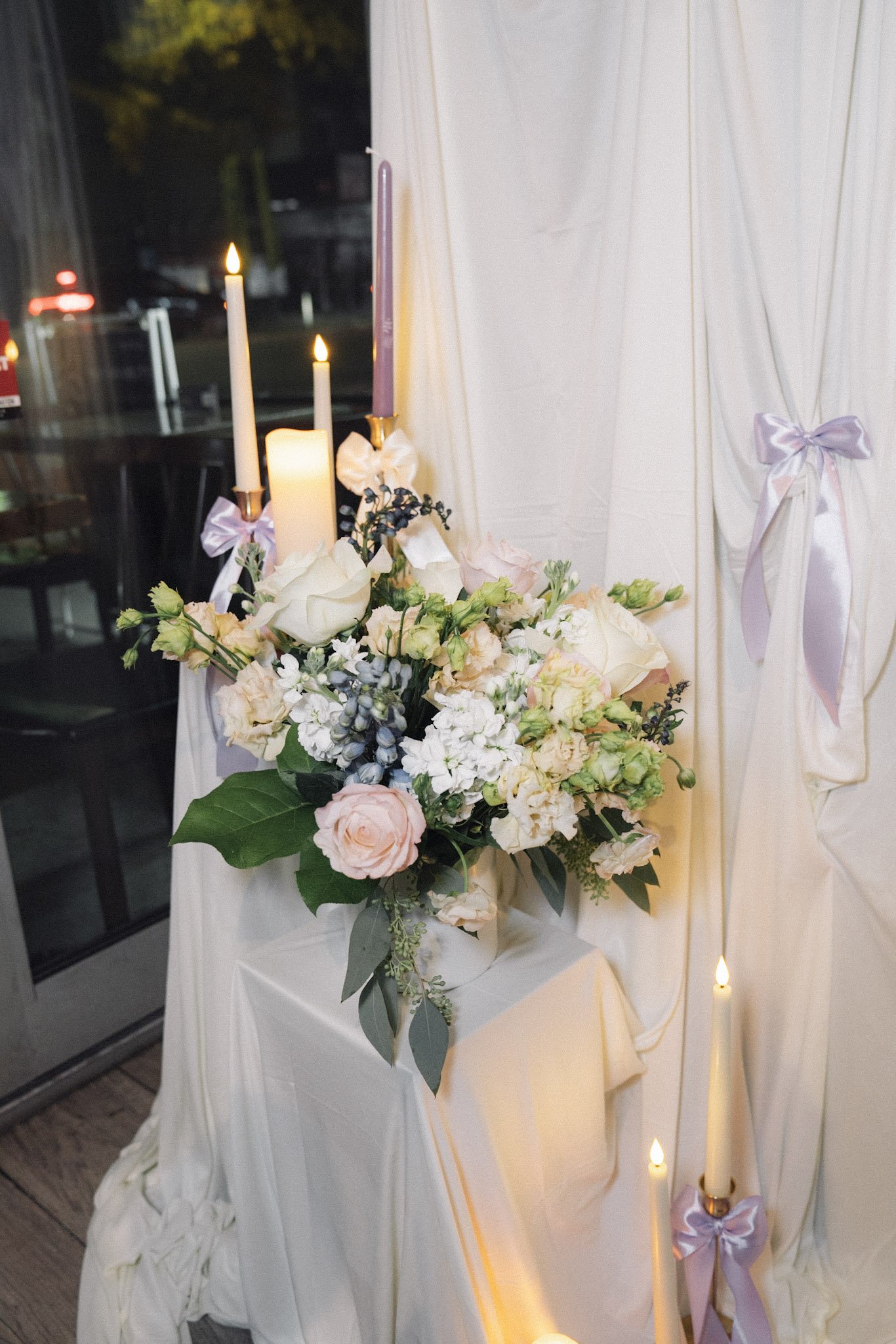 A floral arrangement with pink roses, white flowers, and green leaves on a decorated table with white fabric and candles.