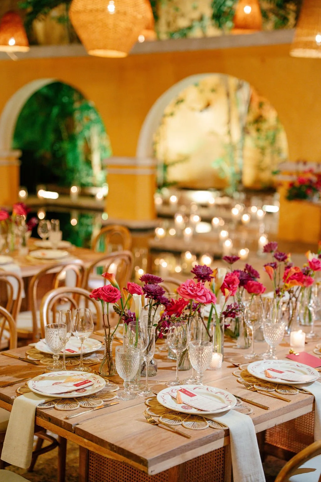 A decorated banquet table with pink and purple flowers, gold and white tableware, and candles, set in a warmly lit room with arched windows.