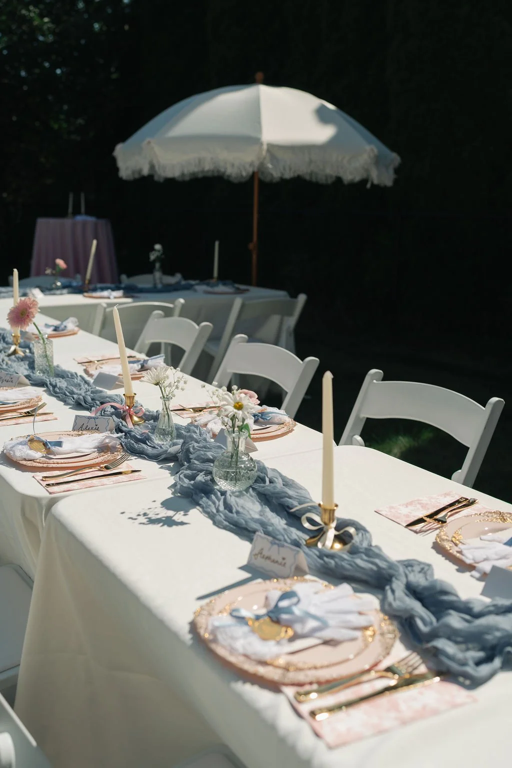 Elegant outdoor dining table set with white tablecloth, floral centerpieces, gold cutlery, pink plates, and tall tapers under a white parasol, with a dark background.