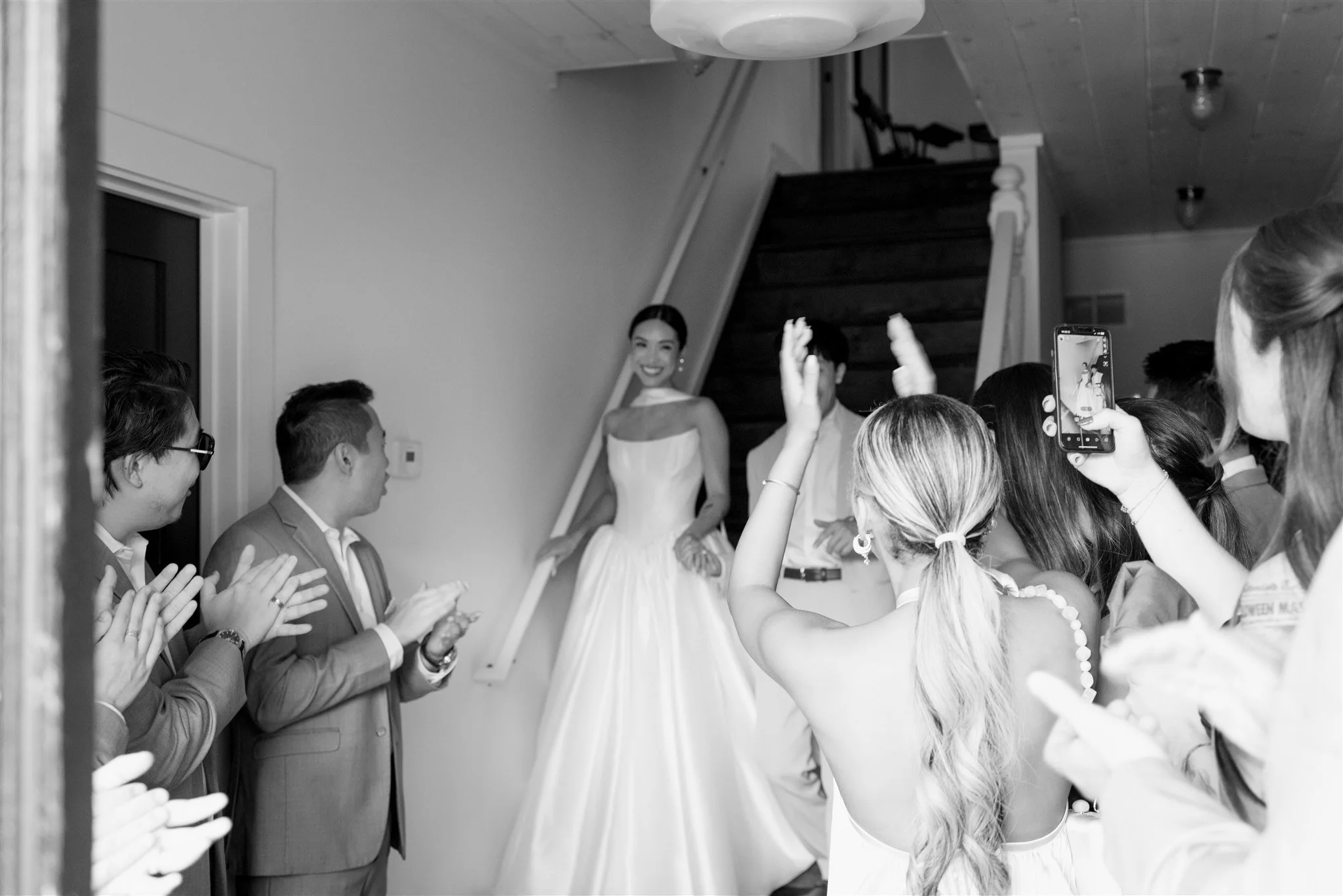 A smiling woman in a dress standing on stairs, surrounded by a group of people who are clapping and taking photos during a celebration or event.