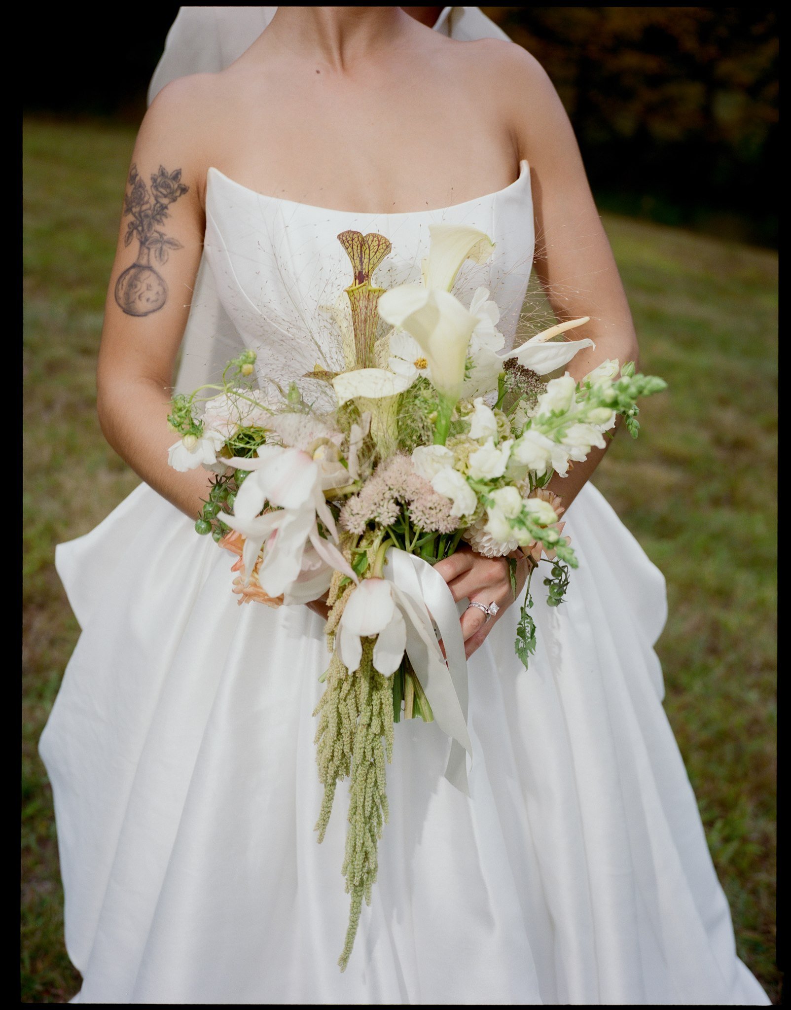 A bride in a white strapless wedding dress holding a bouquet of white and green flowers outdoors, with tattoos on her left arm.