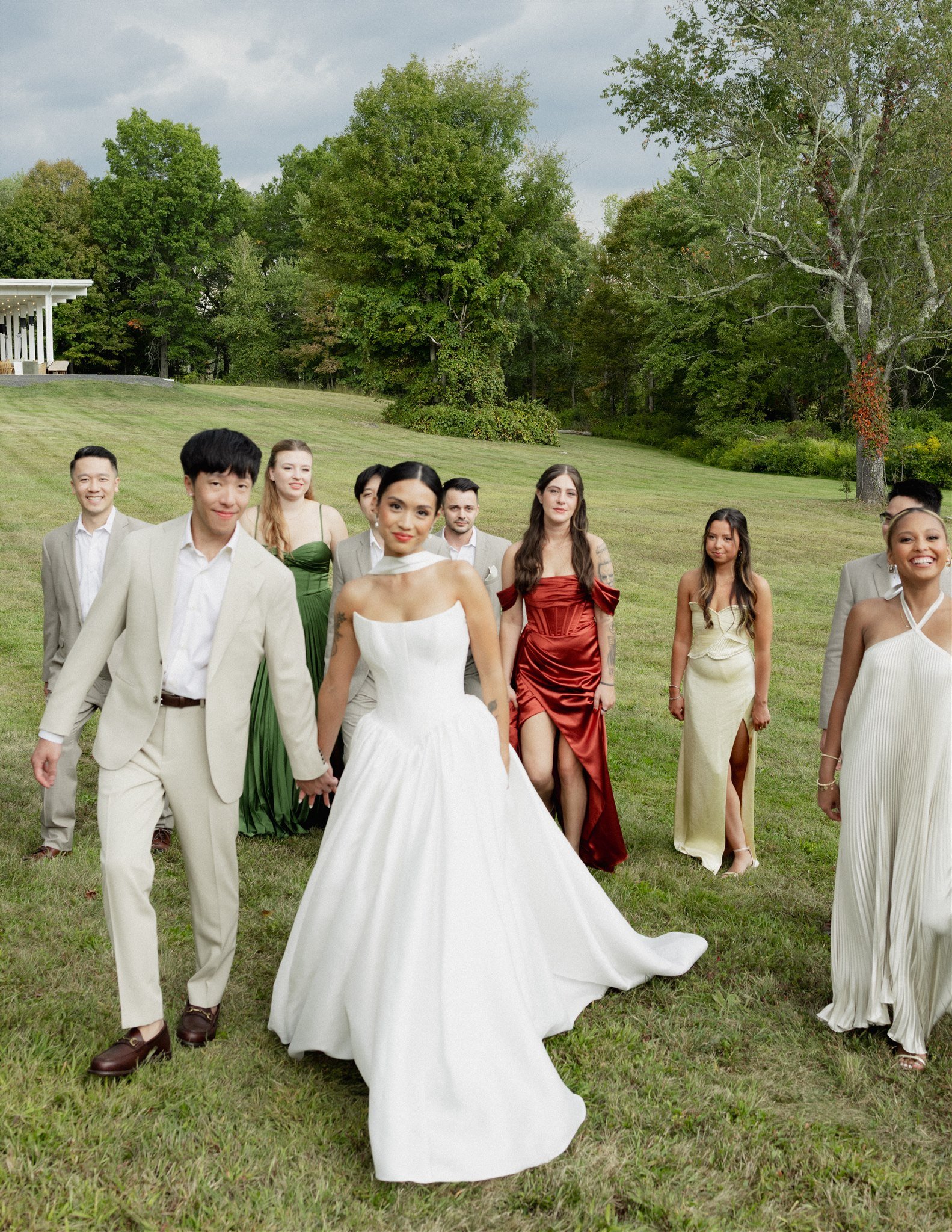 Group of people in formal attire walking on a grassy lawn, with trees and a cloudy sky in the background.