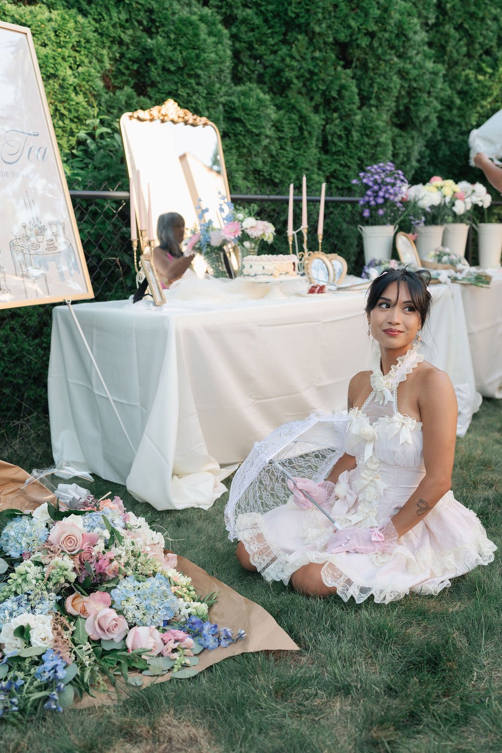 A woman dressed in a vintage wedding dress with lace gloves sitting on the grass, holding a lace parasol, near a table decorated with flowers and cakes at an outdoor garden wedding.