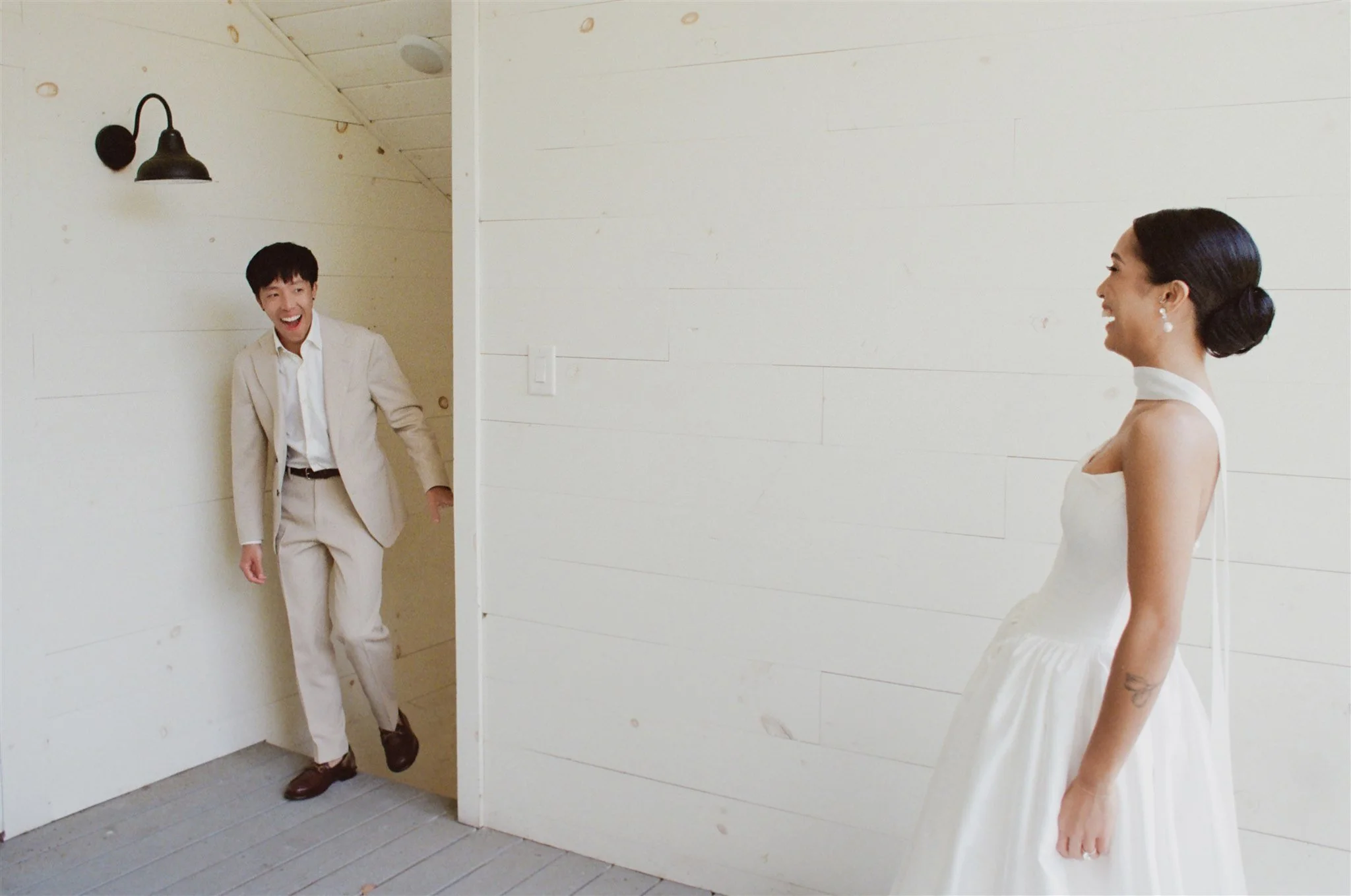 A man in a beige suit peeking out from behind a partial wall and smiling, while a woman in a white dress stands nearby, also smiling at him, in a minimalist room with white wooden panel walls.