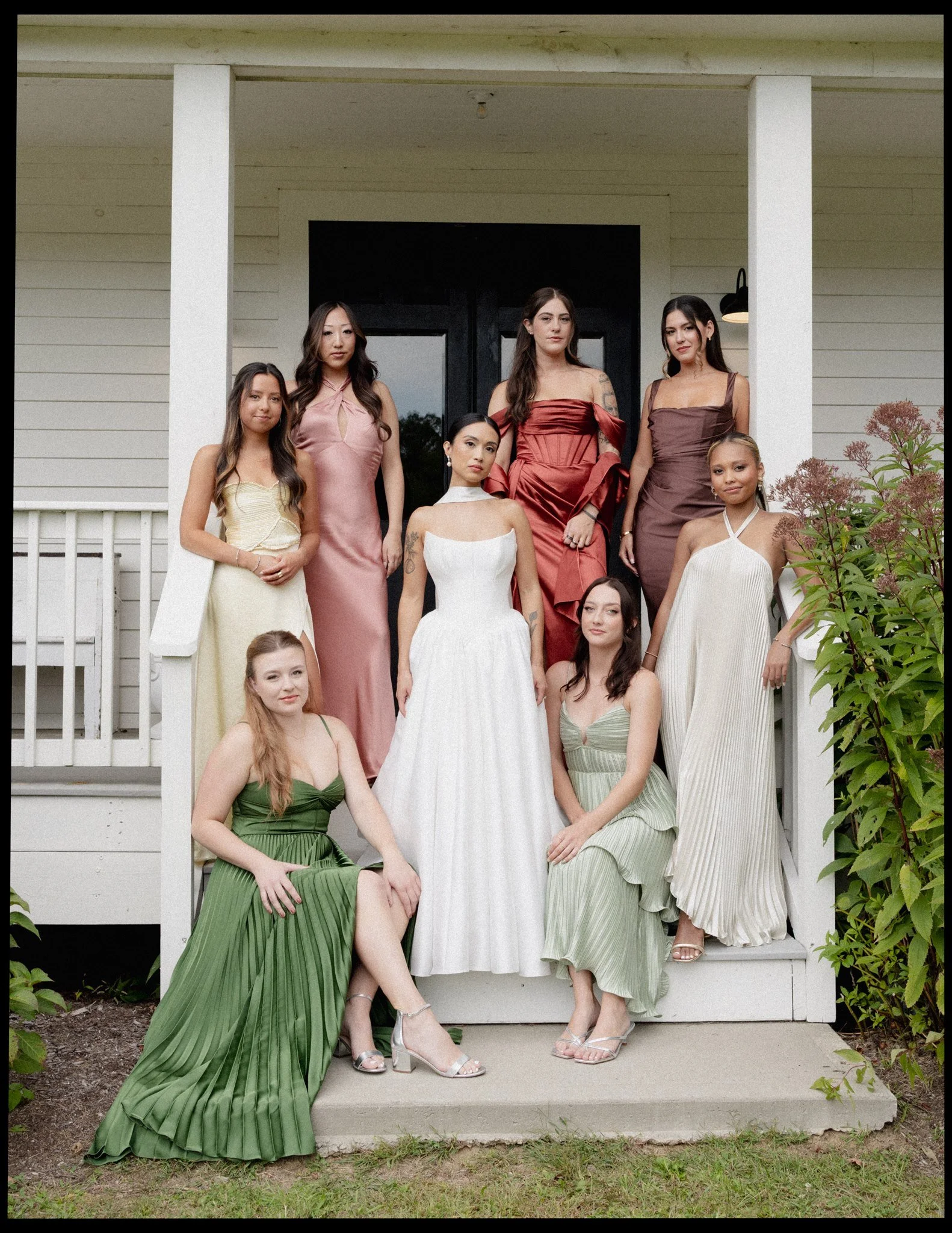 Group of seven women dressed in formal gowns posing on the front porch of a house with white siding and black door, one woman seated on the steps, four women standing behind her, and two women seated on the ground in front, surrounded by greenery.