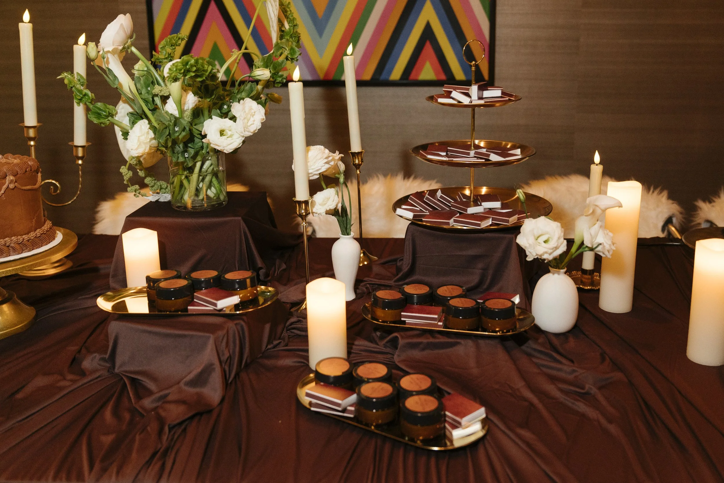 A decorated table with candles, flowers, chocolates, and a cake, set against a colorful geometric art background.
