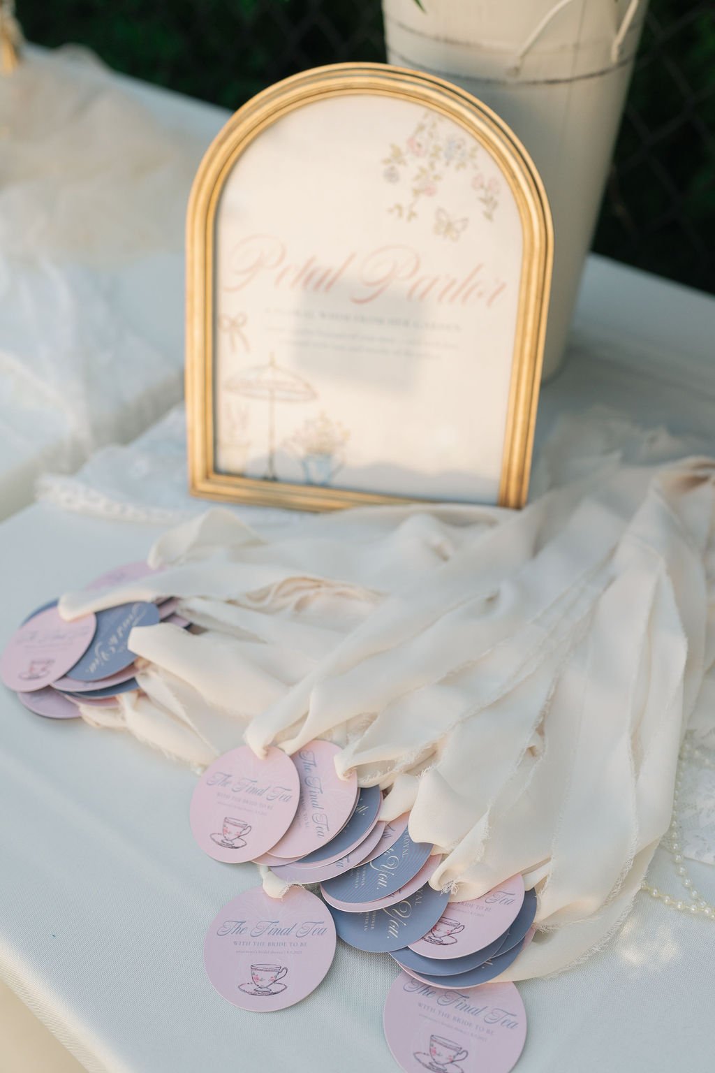 A framed sign with elegant script and pastel colors, reading 'Hello Party'. A table holding pink and purple circular tags with the text 'The Final Tea' and a teacup illustration, draped with light fabric or ribbons.
