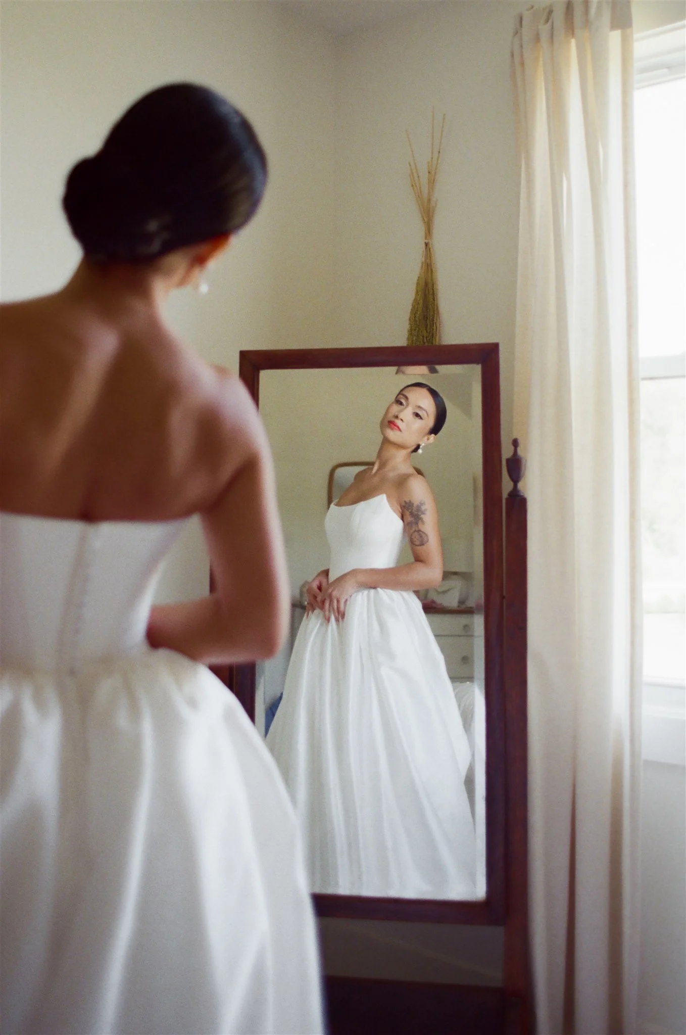 A woman in a white dress looking at herself in a mirror in a bedroom with cream-colored curtains.