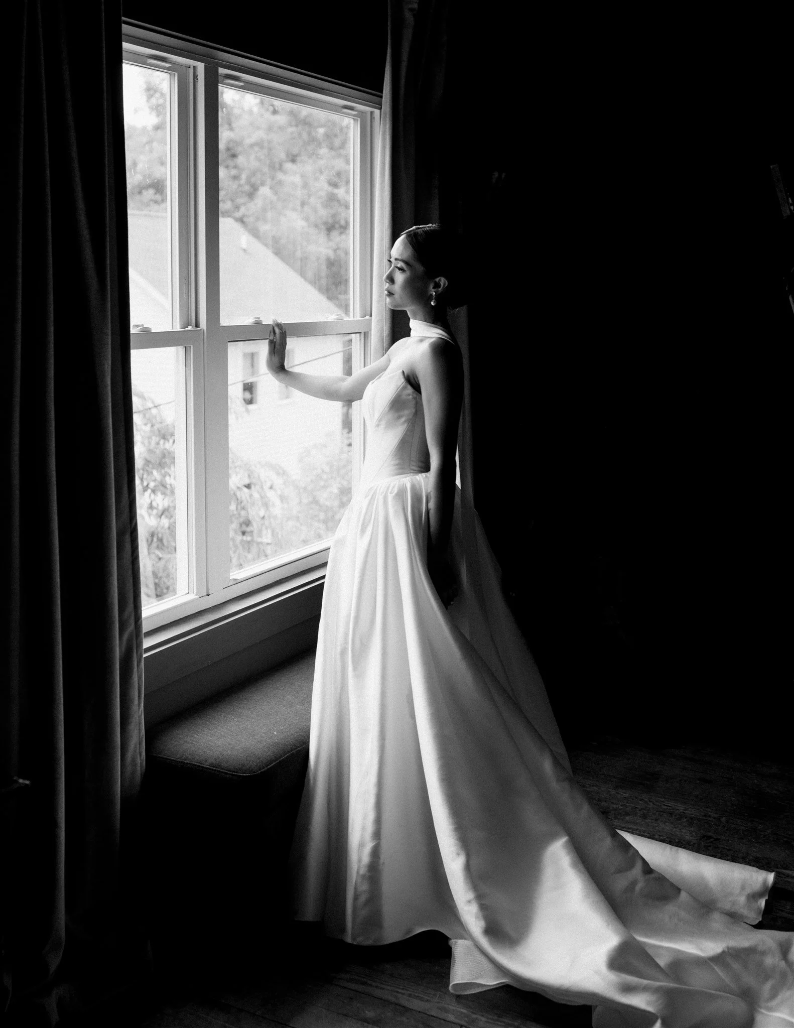A woman in a wedding dress looking out a window with a view of trees, with her hand resting on the window frame, in black and white indoor setting.