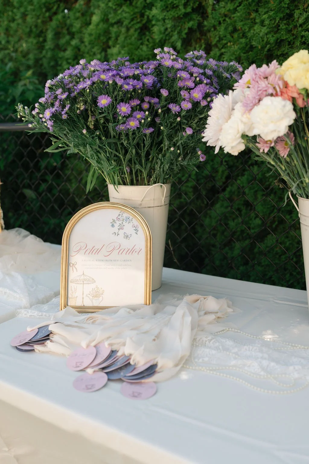 Purple and pink flowers in white vases at a floral display set on a table with a sign that reads 'Petal Parlor' and various fabric and pearl accessories.