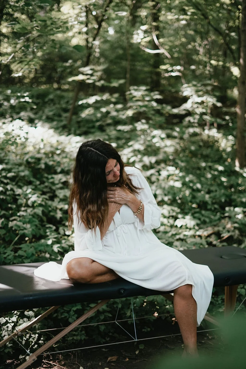 A woman in a white dress sitting on a massage table in a forest, touching her chest with a peaceful expression. . Experiencing healing through somatic coaching.