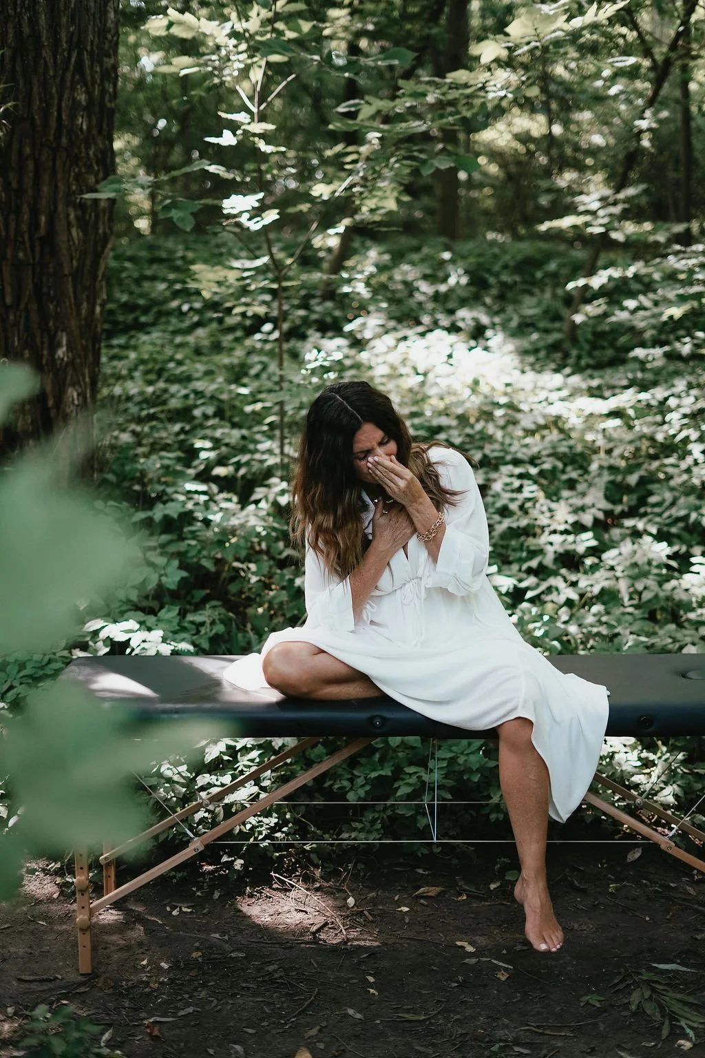 Woman sitting on a massage table outdoors in a forested area, covering her face with one hand and clutching her chest with the other, wearing a white dress.. Experiencing healing through somatic coaching.