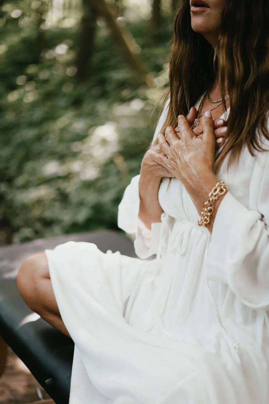 A woman sitting outdoors with her hands over her chest, wearing a white dress and gold jewelry, in a natural setting with green trees and sunlight in the background. . Experiencing healing through somatic coaching.