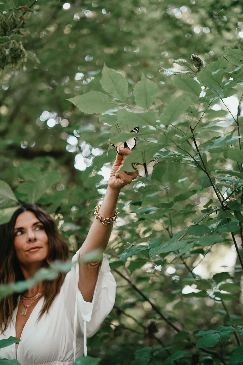 Woman with wavy hair and gold jewelry reaching up among green leaves, surrounded by butterflies in a wooded area. . Experiencing healing through somatic coaching.