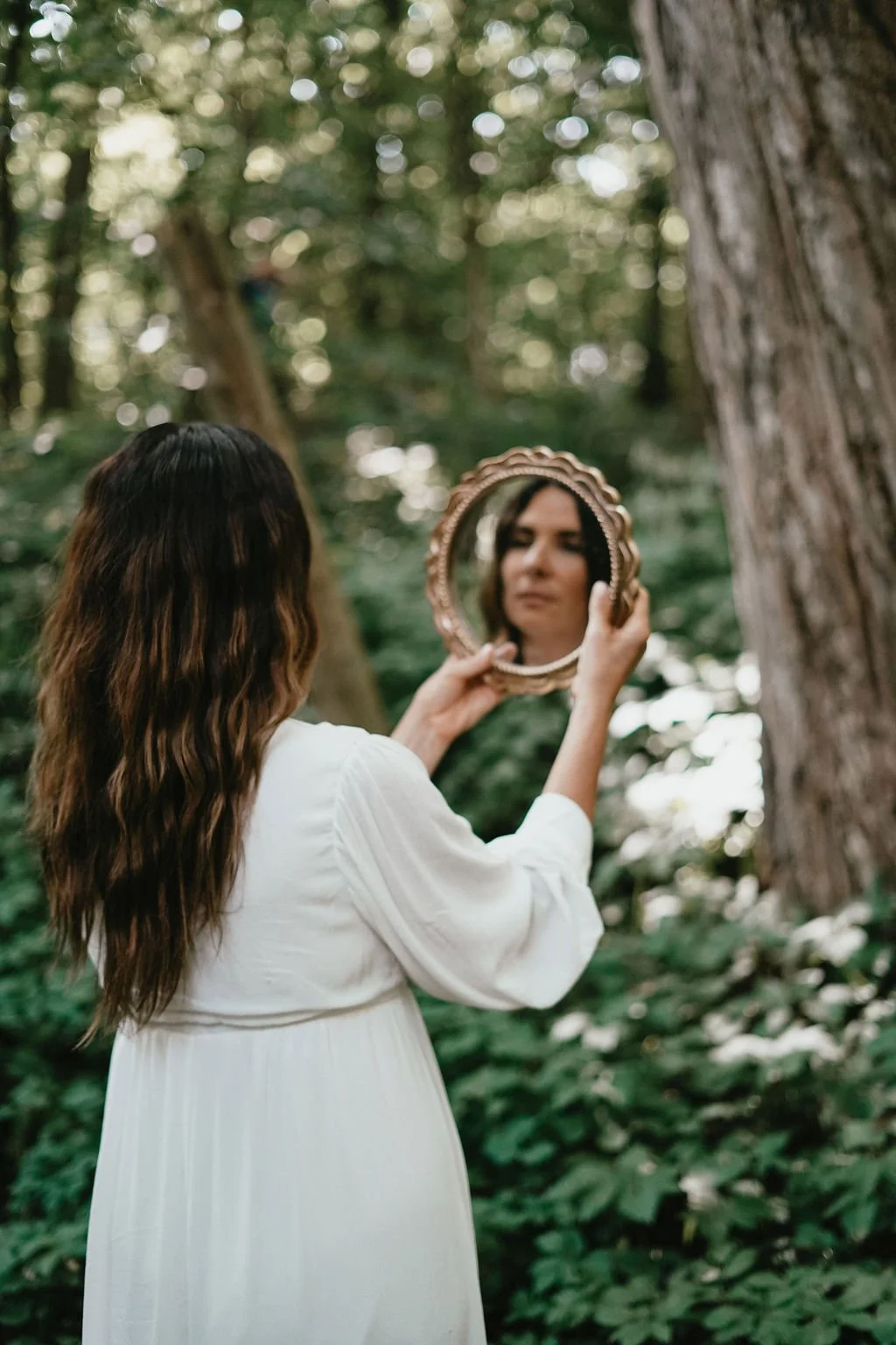 A woman with long wavy hair wearing a white dress looks into a mirror she is holding in a forest surrounded by green trees and bushes. . Experiencing healing through somatic coaching.