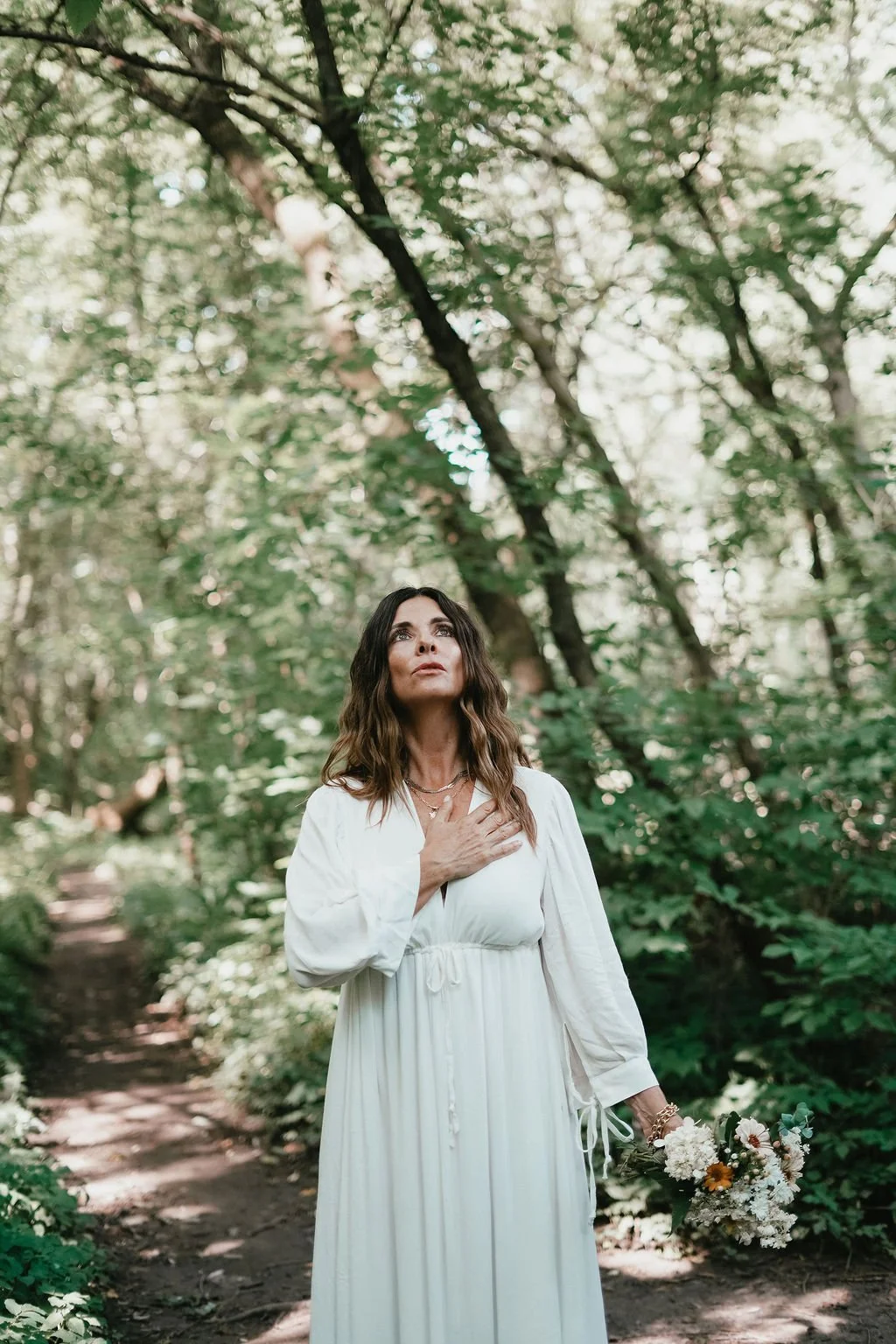 A woman in a flowing white dress stands on a forest trail, holding a bouquet of flowers in her left hand and placing her right hand on her chest, looking upward with a serene expression. . Experiencing healing through somatic coach
