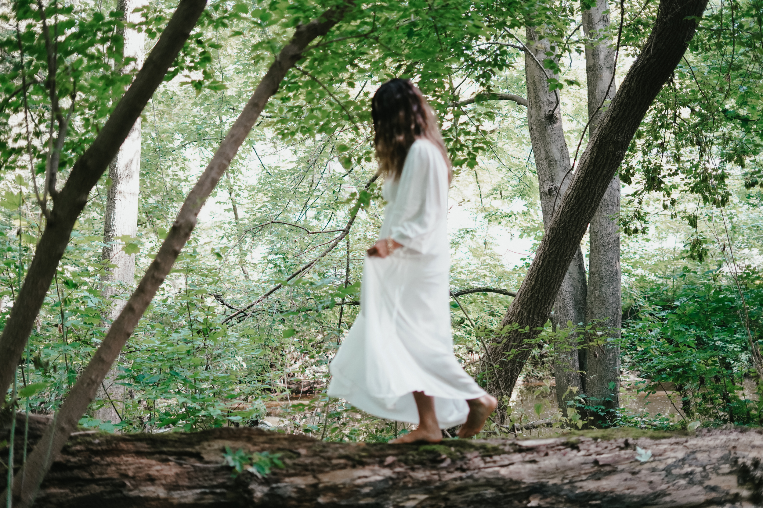 Woman in a white dress walking on a fallen tree in a green forest. . Experiencing healing through somatic coaching.