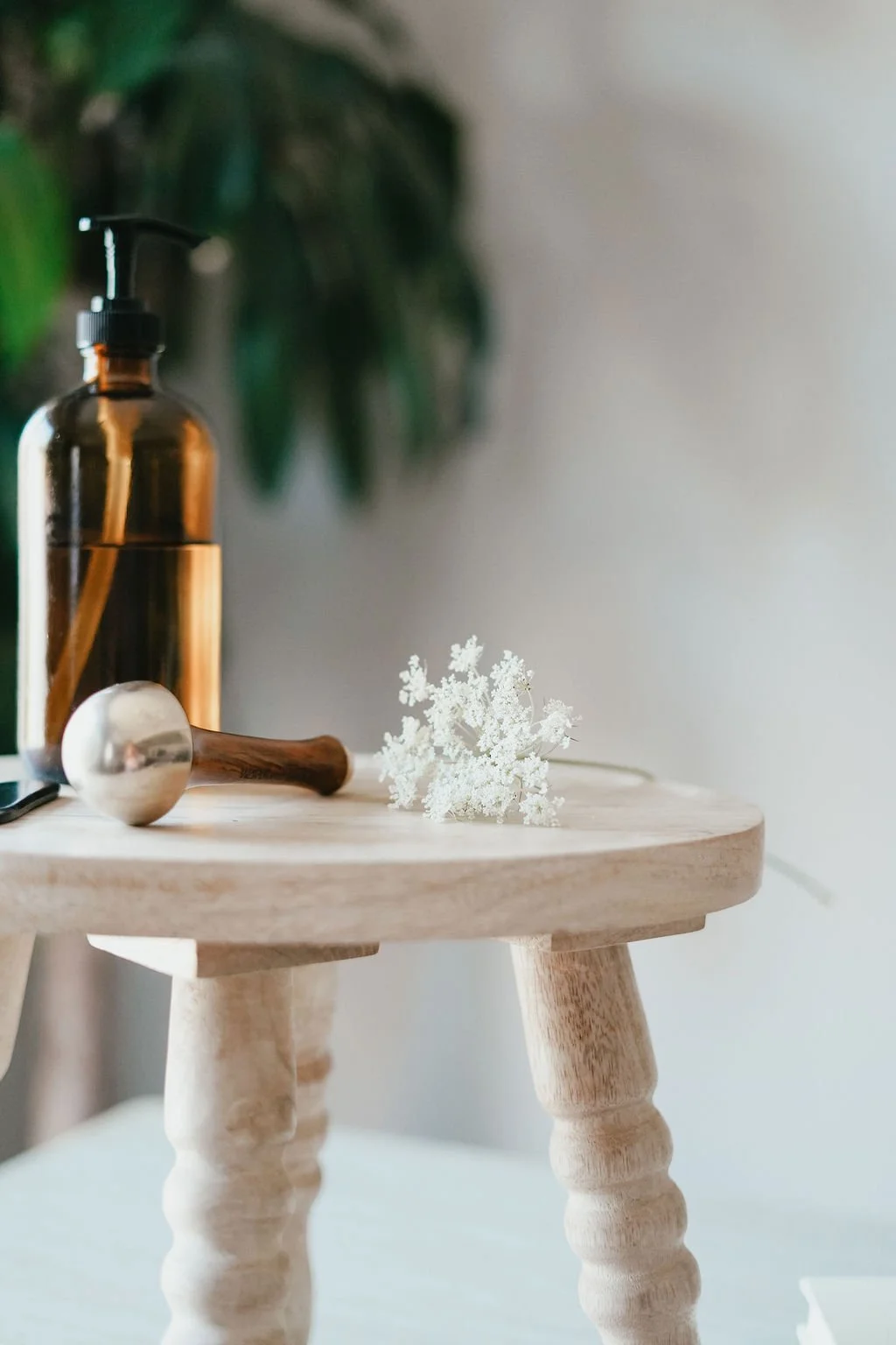 A wooden table with a glass pump bottle, a metal massage hammer, and a small white decorative sculpture on top. Green leaves are blurred in the background. Booking a glow state facial in stratford ontario.
