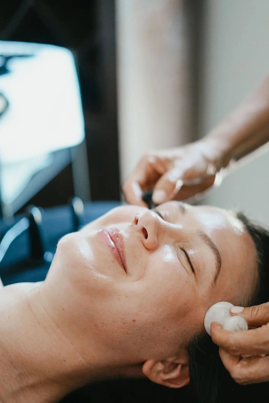 A woman enjoying a facial treatment, lying down with her eyes closed, as a skincare professional applies a product to her face. The setting appears to be a spa or skincare clinic.